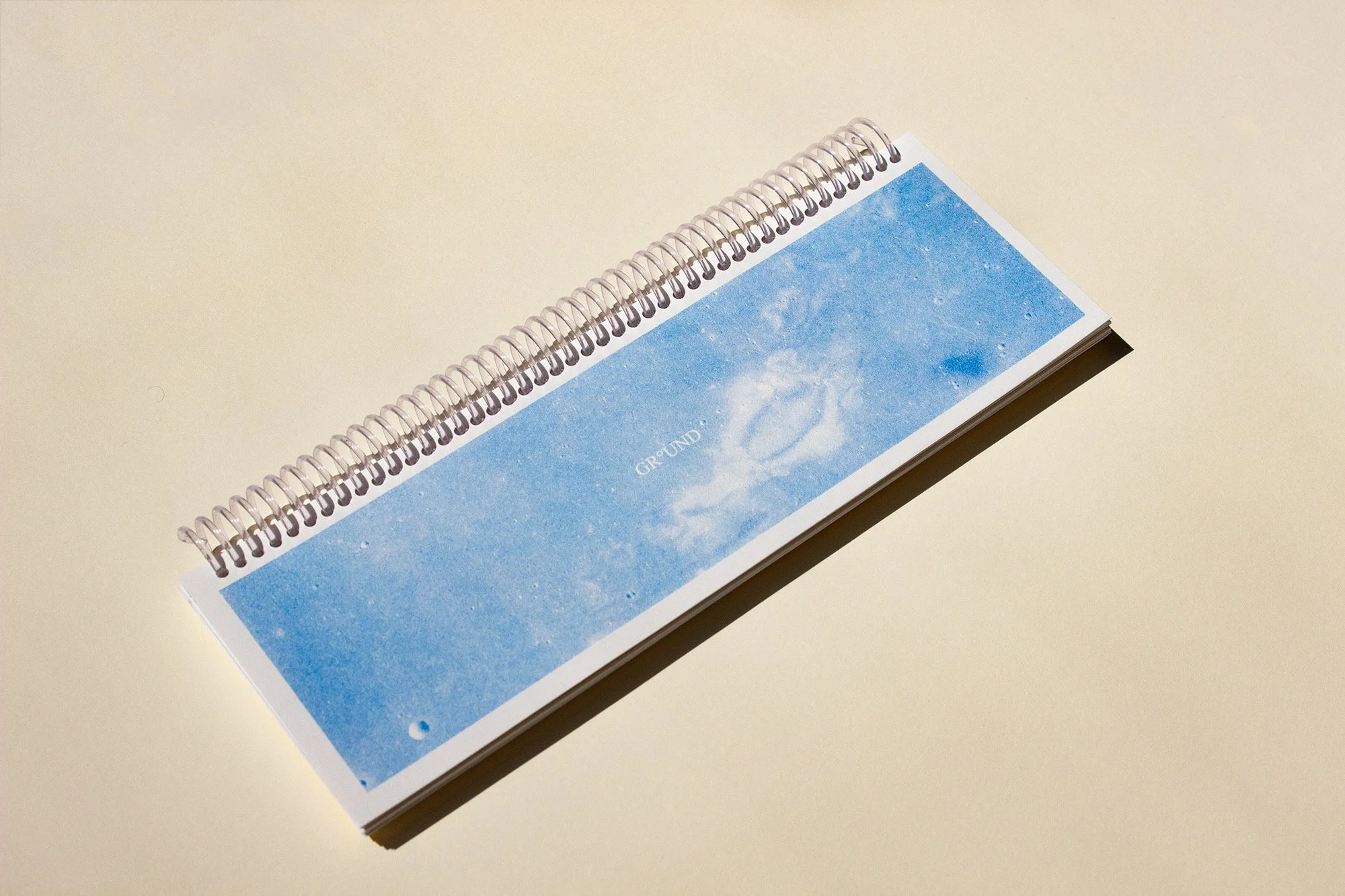 A spiral-bound notebook with a cover image of a blue sky and moon surface, lying on a beige surface. Document is the print version of GROUND, an art-RPG published by UKAI Projects