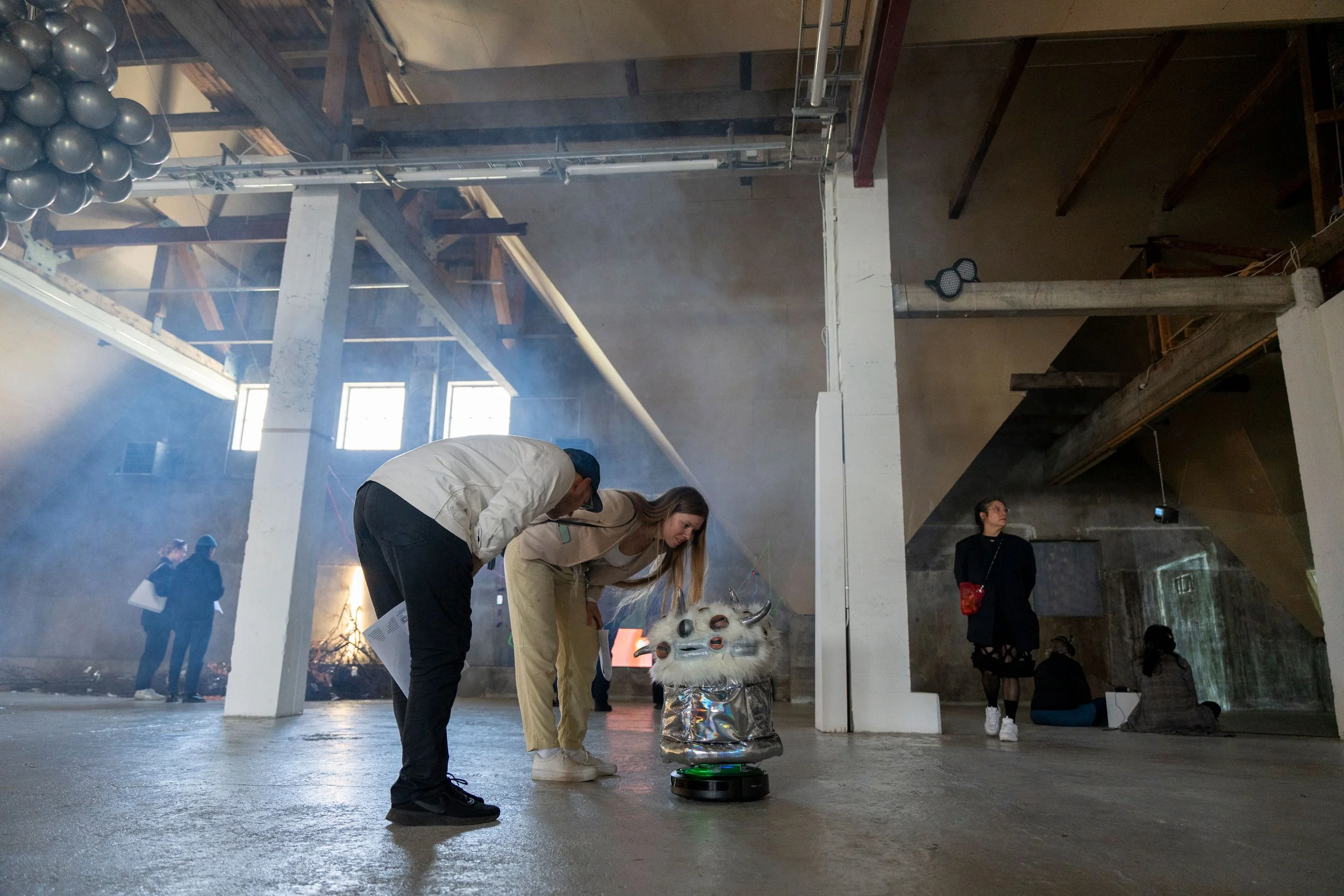 People in an industrial-style room engaged in an activity with a lion dance head. The room has exposed beams, scaffolding, and a foggy atmosphere. Several individuals are observing or participating in the event, with some wearing dark clothing.