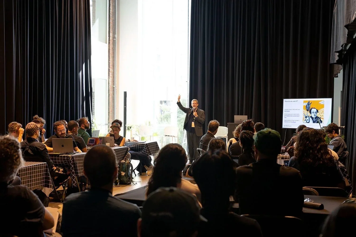 A man giving a presentation at an indoor conference with attendees seated around tables, some taking notes or working on laptops. The presenter is standing near a screen with a slide, and large windows let in natural light.