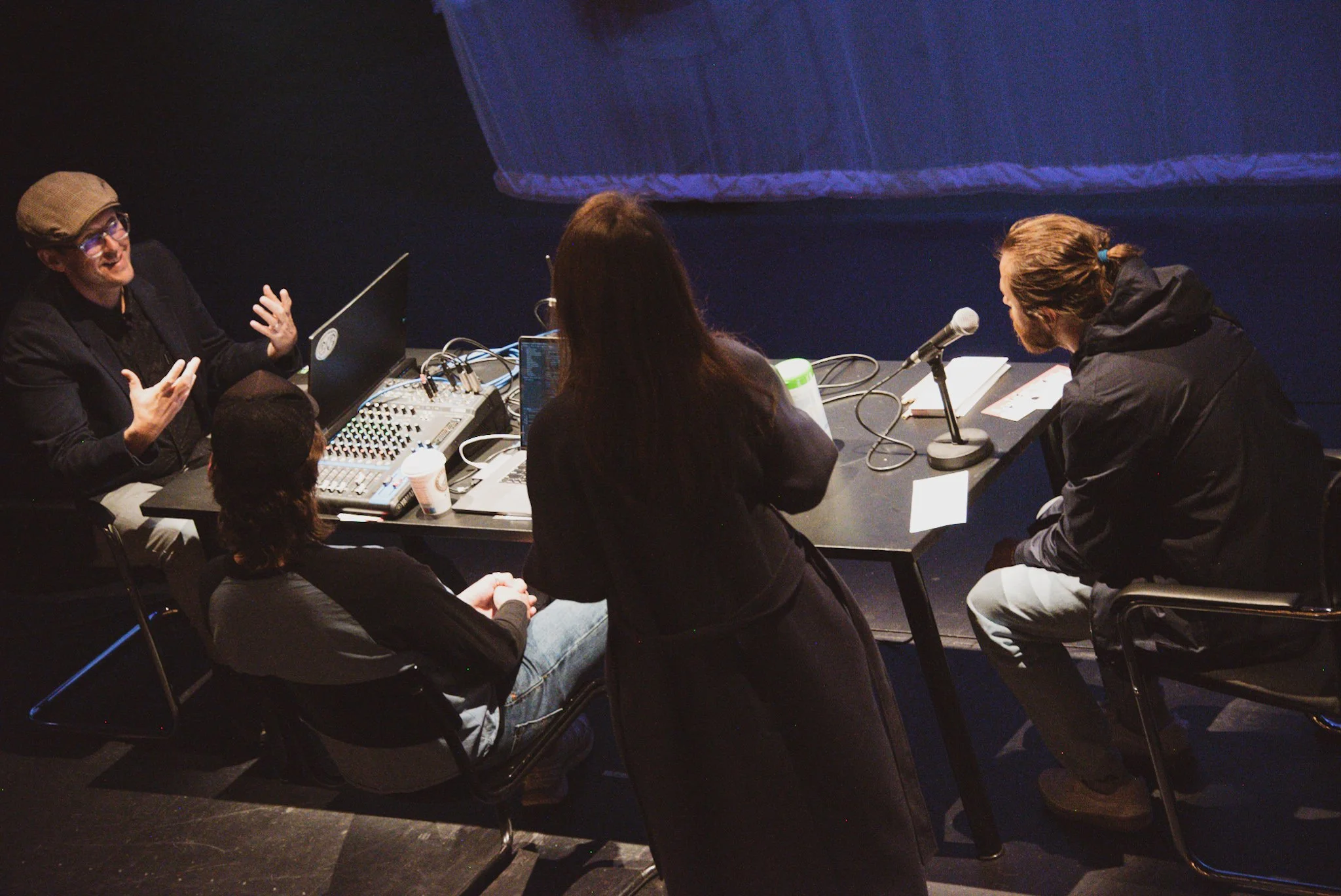 People sitting around a table in a dark room during a podcast or radio recording, with audio equipment and a microphone visible, engaged in conversation.