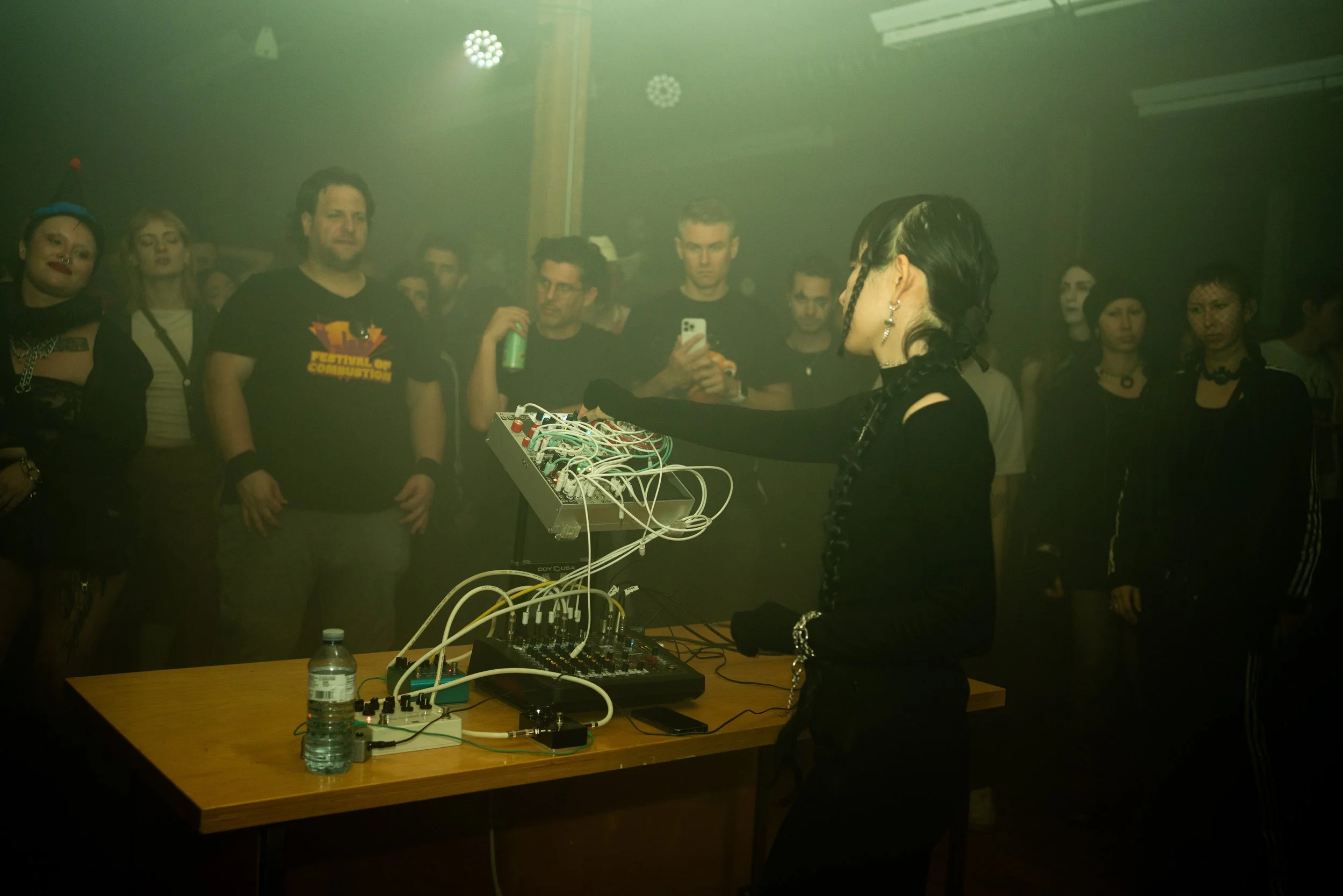A woman with black braided hair performing with electronic music equipment in front of a crowd in a dark, smoky room. The audience includes men and women, some holding drinks and one person taking a photo.