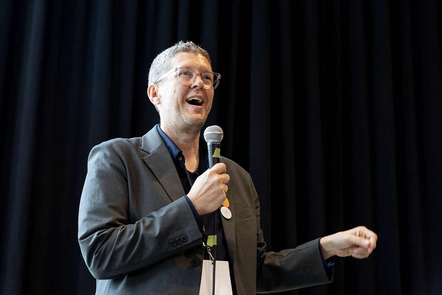 A man speaking into a microphone during a presentation or speech, standing in front of a dark curtain.