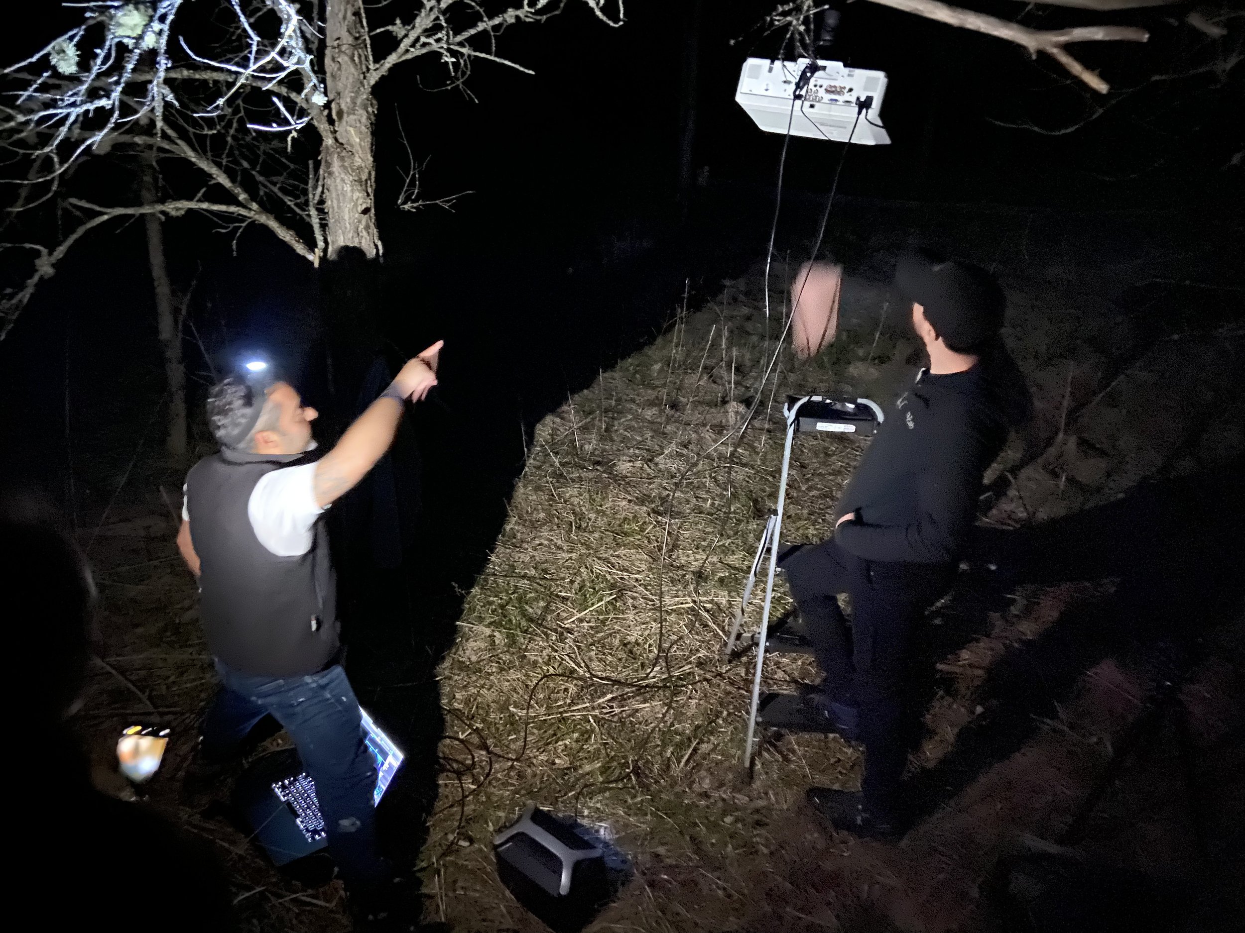 Nighttime outdoor scene with three individuals, two men and one woman, gathered around electronic equipment, including laptops and a large white device hanging from a tree. One person is pointing at something in the tree while the others observe. The area is illuminated by headlamps and the equipment's glow.