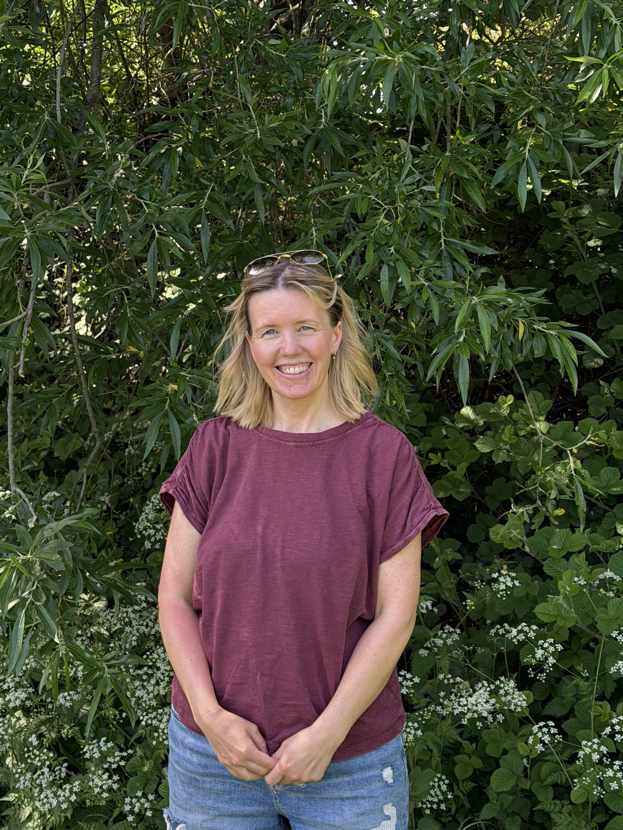 A woman with blonde hair smiling outdoors in front of green leafy bushes, wearing sunglasses on her head, a maroon T-shirt, and ripped blue jeans.