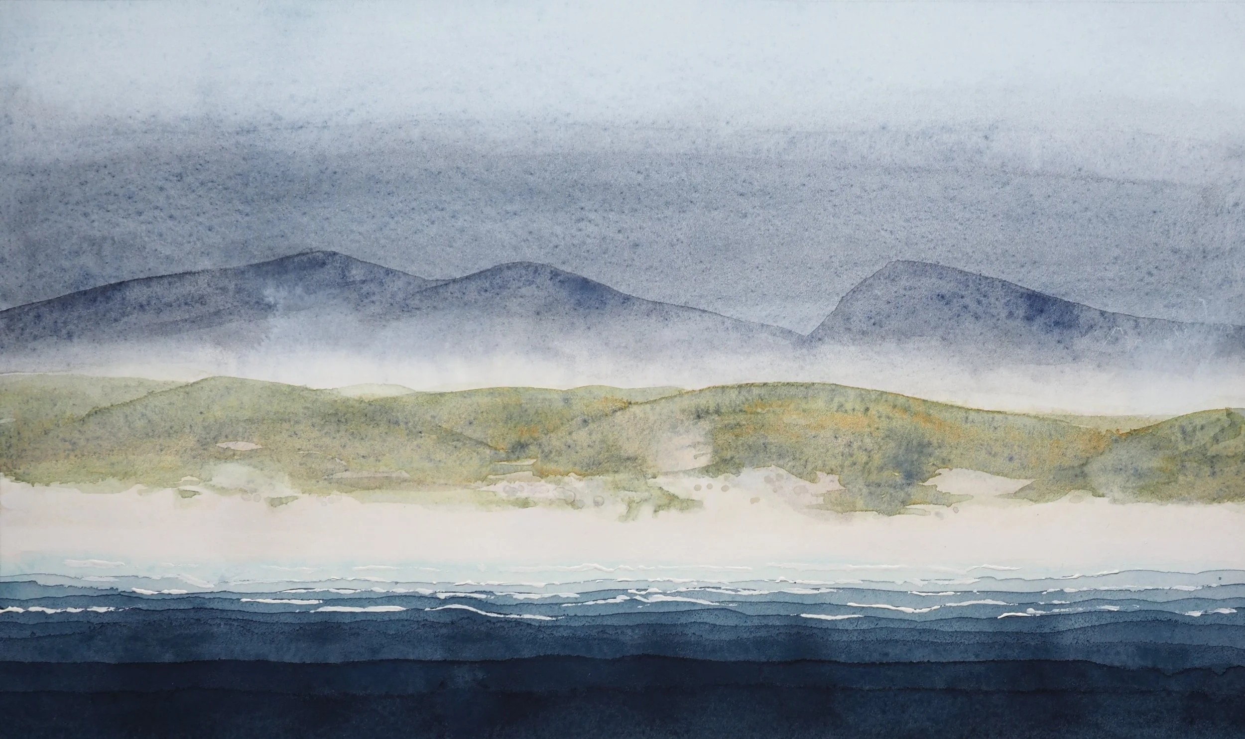 Deep blue water brake on a shell sand beach. Beyond are misty sand dunes with grey-blue hills in the distance.
