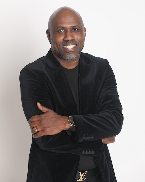Man with a bald head and beard smiling, wearing a black blazer and black shirt, standing against a plain white background.