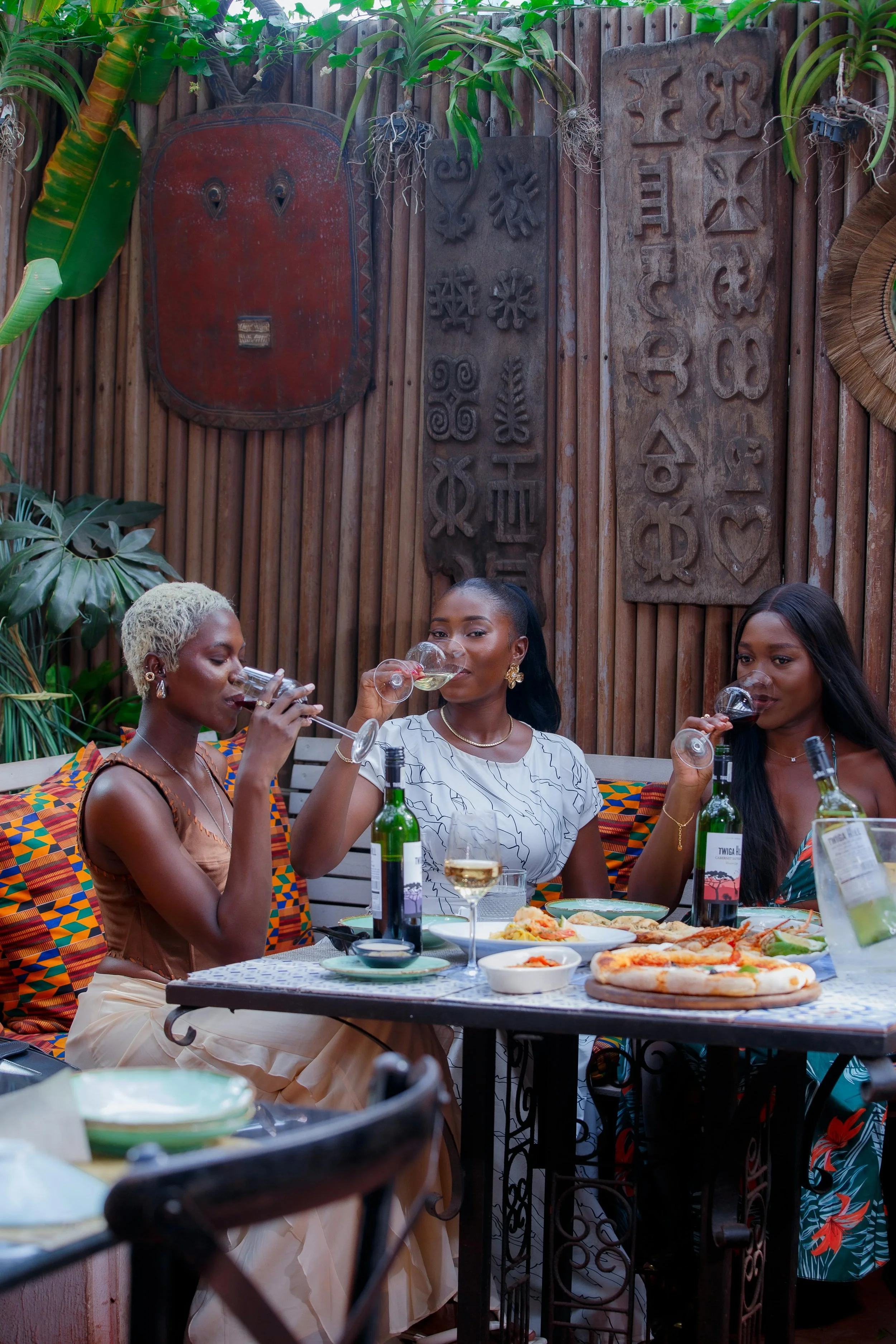 Three women sitting at a table enjoying drinks and food in an outdoor setting decorated with African art and lush green plants.