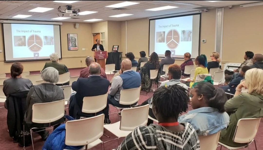 A speaker giving a presentation titled 'The Impact of Trauma' to an audience in a conference room with two large screens displaying the presentation slides.
