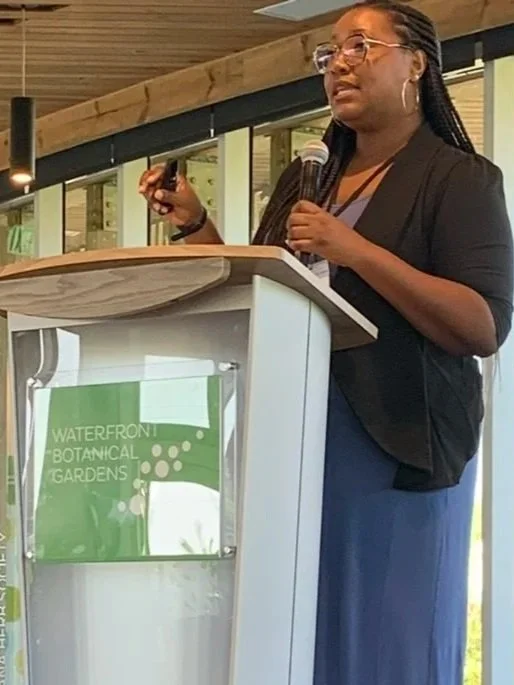 Woman speaking at a podium during an event at Waterfront Botanical Gardens, holding a microphone and a remote control.