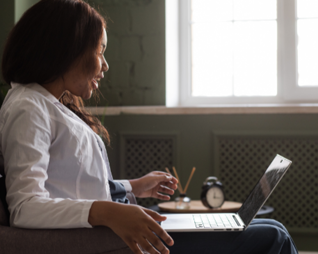 A woman with long reddish-brown hair sitting on a gray couch, typing on a laptop with her right hand and holding a coffee mug with her left, next to a small round clock and a small container with wooden sticks, in front of a window with sunlight streaming in.