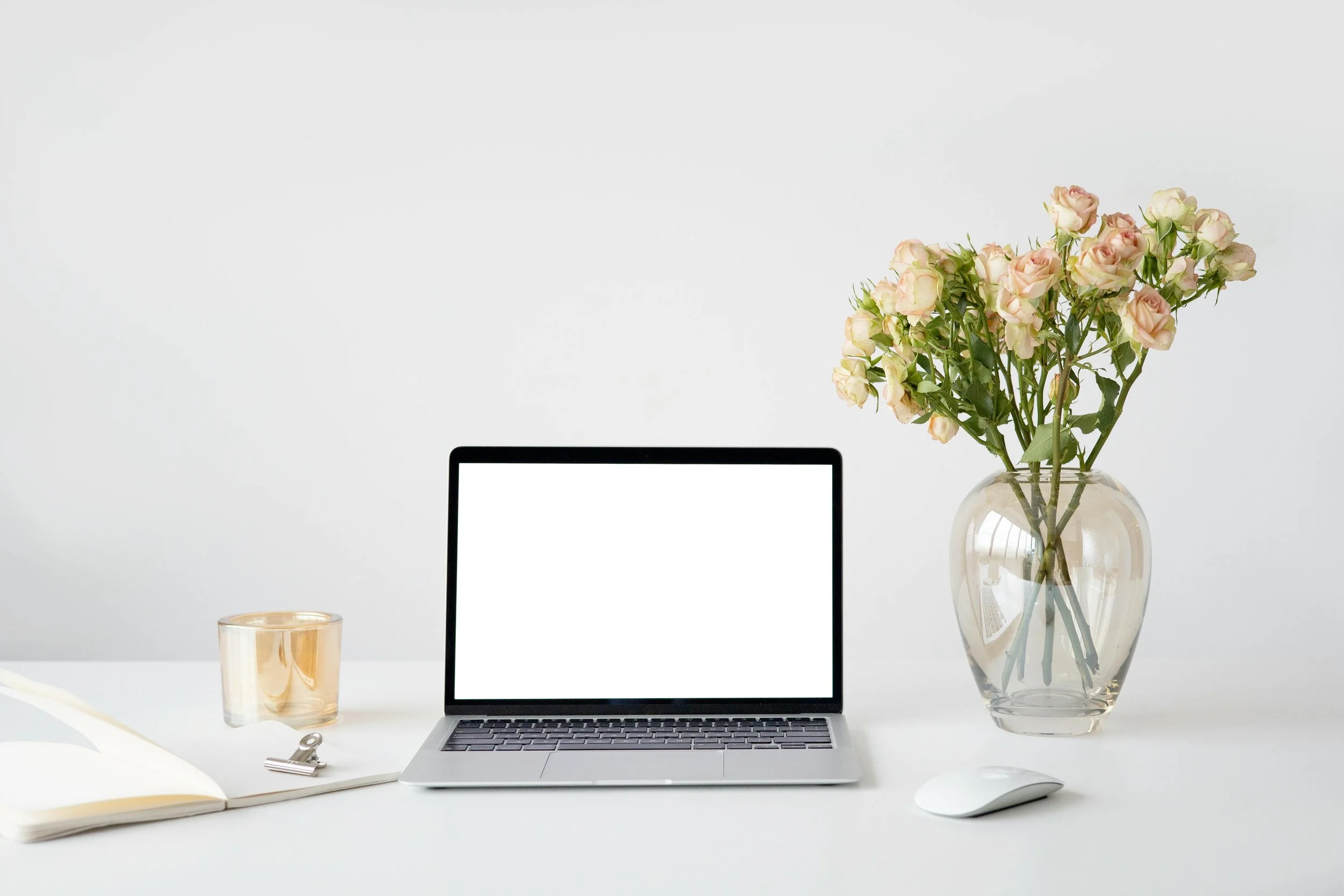 A workspace with a silver laptop, a white mouse, an open notebook with a pen, a glass of water, a large clear vase with pink and white roses, and a glass container, all arranged on a white surface against a plain white background.