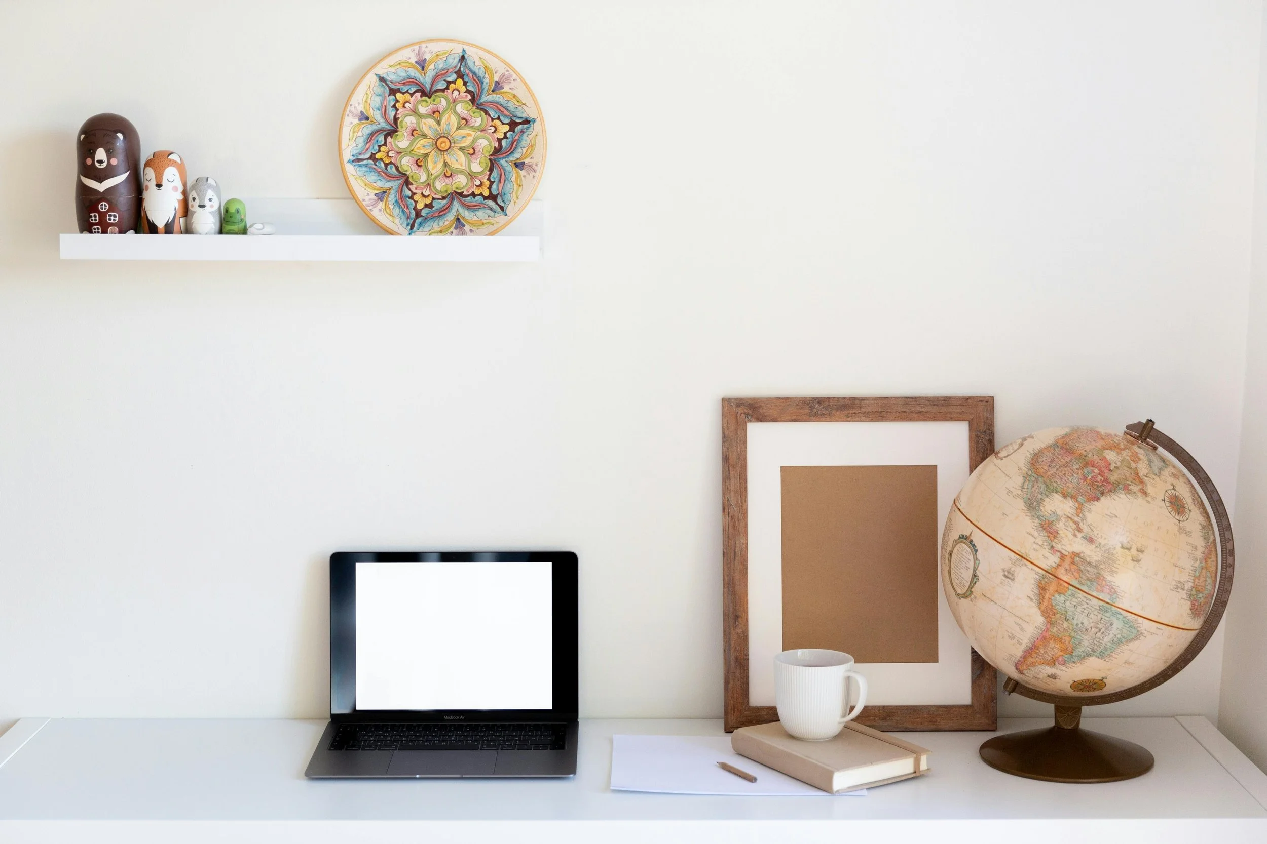 A white desk against a white wall with a laptop, a mug, a notebook, a pen, empty picture frames, a globe, and a floating shelf with decorative items, including painted wooden animal figures and a decorative plate with floral design.