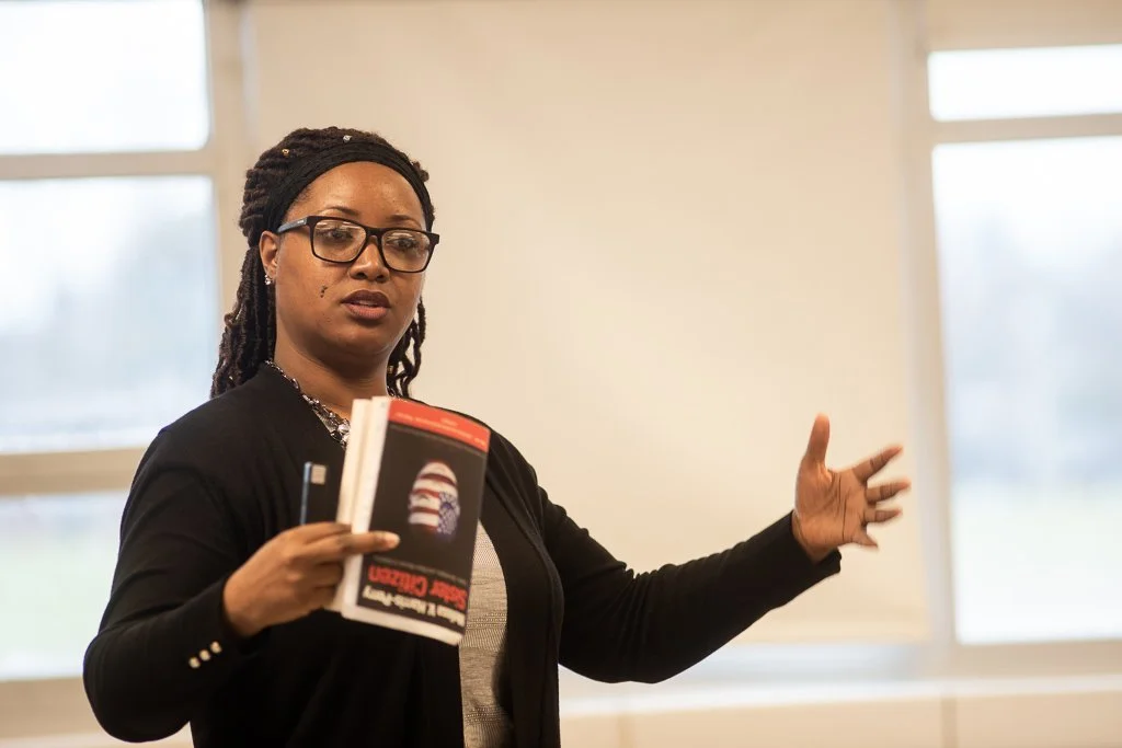 A woman with glasses, dreadlocks, and a black cardigan standing in front of windows, holding a book and gesturing as if giving a presentation.