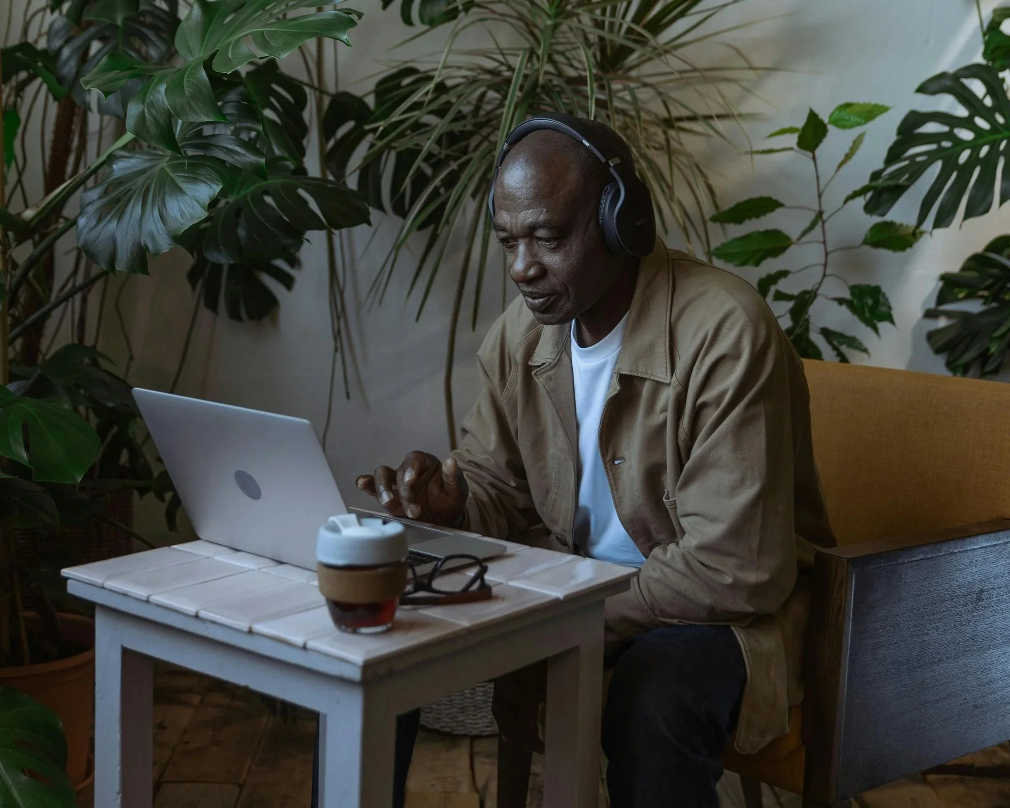 A man wearing headphones and a beige jacket using a laptop at a small white outdoor table surrounded by large green plants.