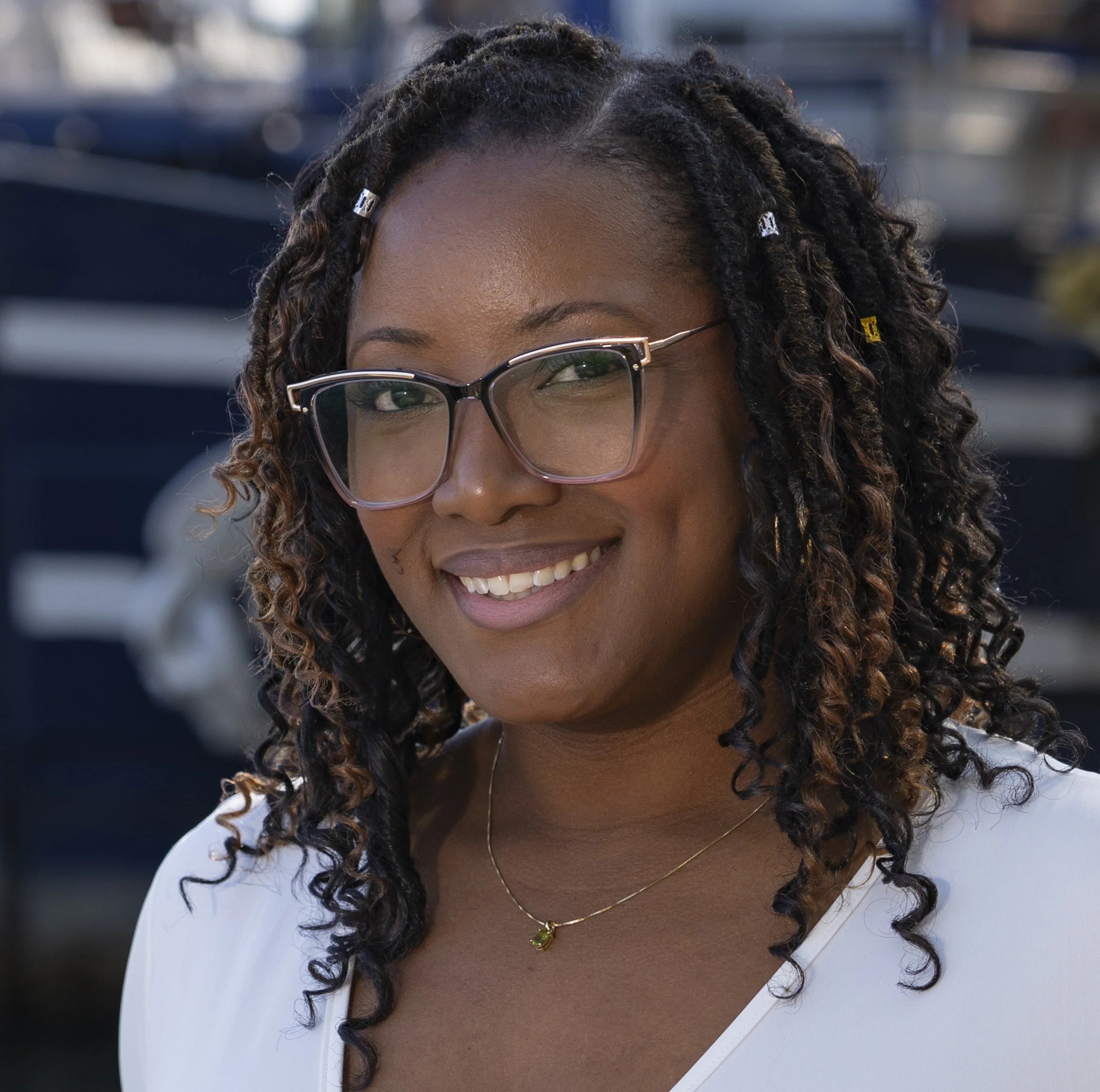 A woman with curly hair and glasses smiling, wearing a white top with a necklace, outdoors with blurred background.