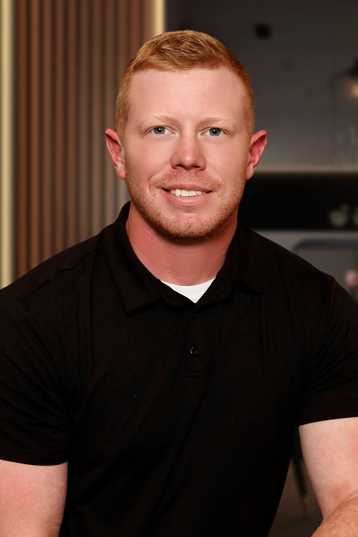 Headshot of a young man with short red hair, blue eyes, and a slight beard, wearing a black shirt, smiling in an indoor setting.