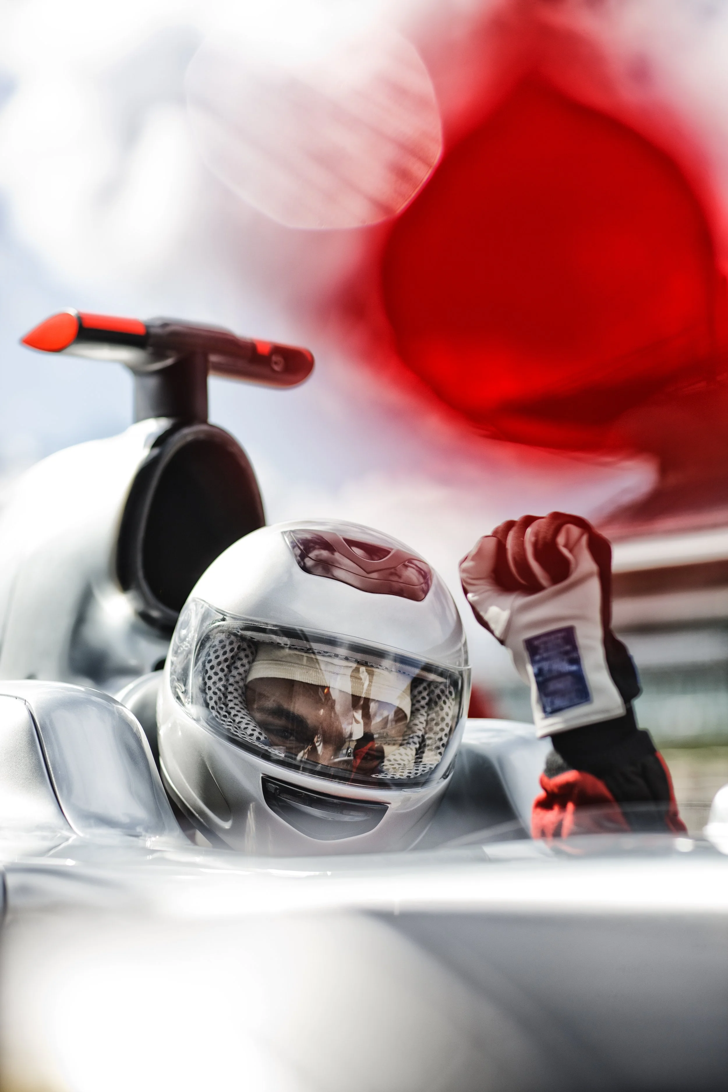 A person wearing a motorcycle helmet with a clear visor and racing suit, standing next to a white motorcycle, with a blurred red and white background.