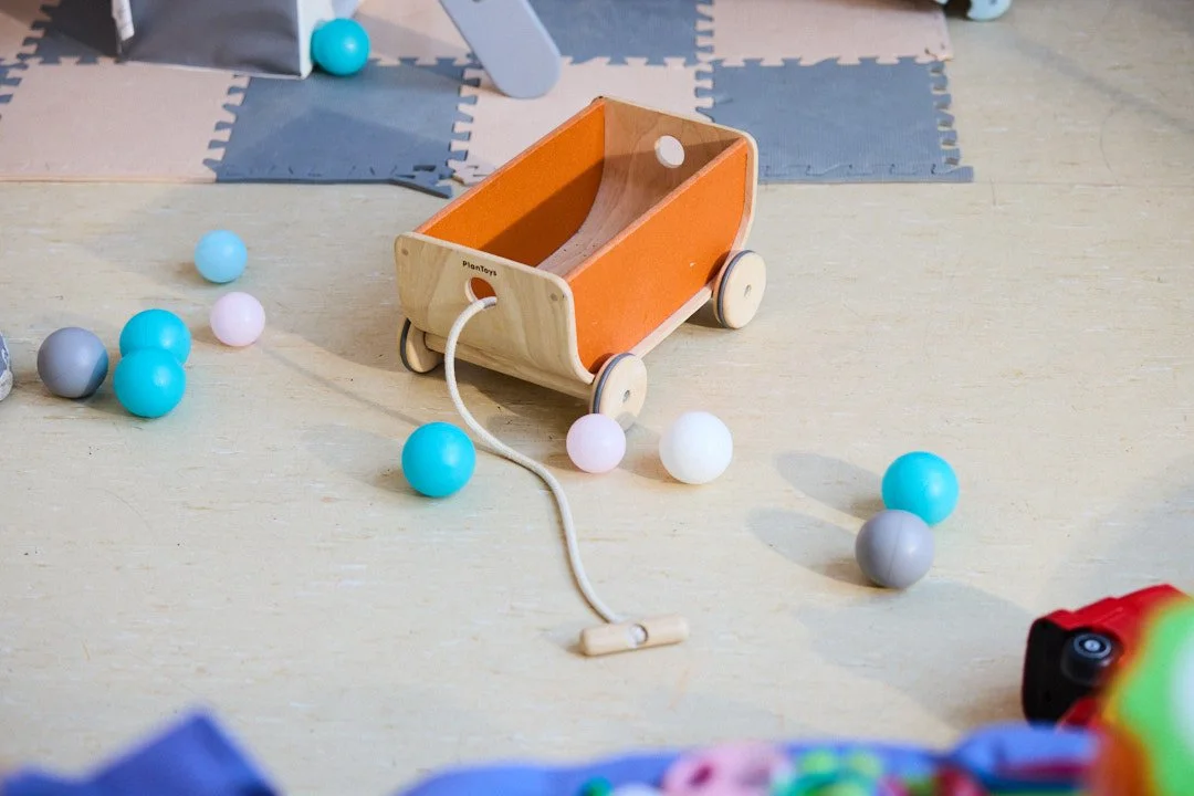 A wooden toy wagon with orange sides, white wheels, and a string attached, is on a light wooden surface surrounded by scattered pastel-colored balls, with foam puzzle mats in the background.