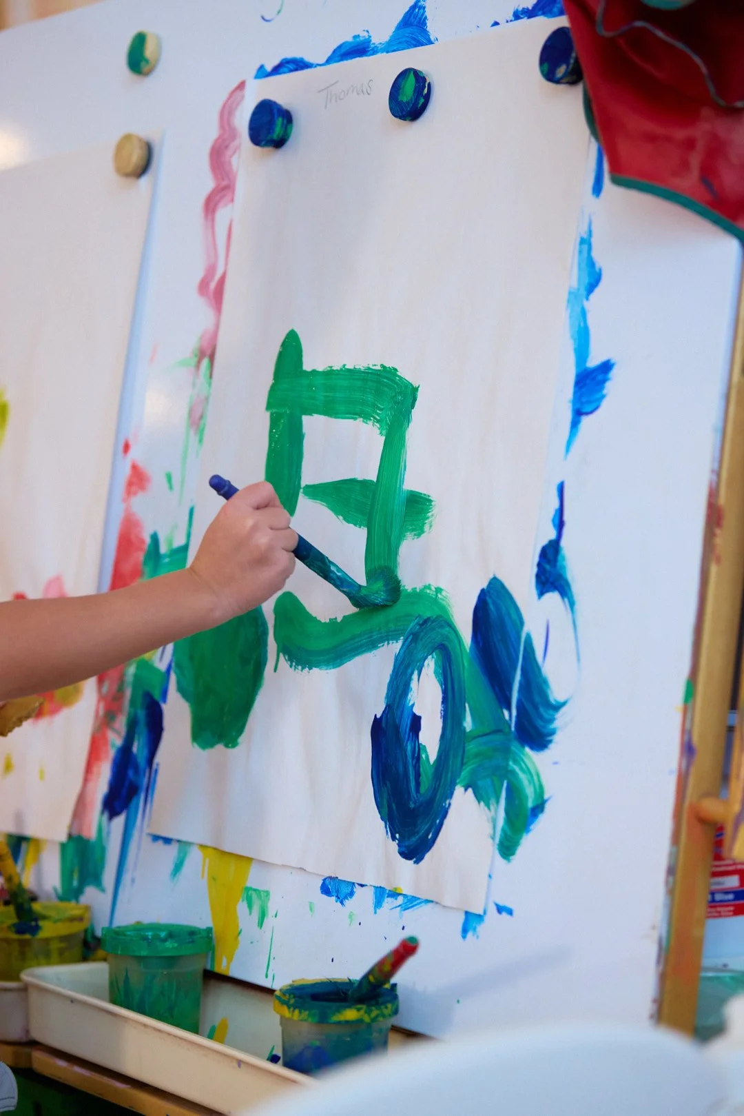 Child's hand painting a green and blue letter 'E' on a white canvas with thumb and brush at an art studio.