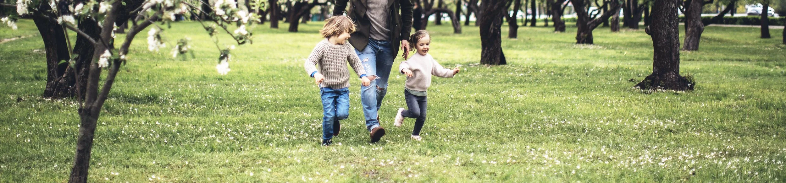 Two children and an adult walking through a park with green grass and trees in bloom.