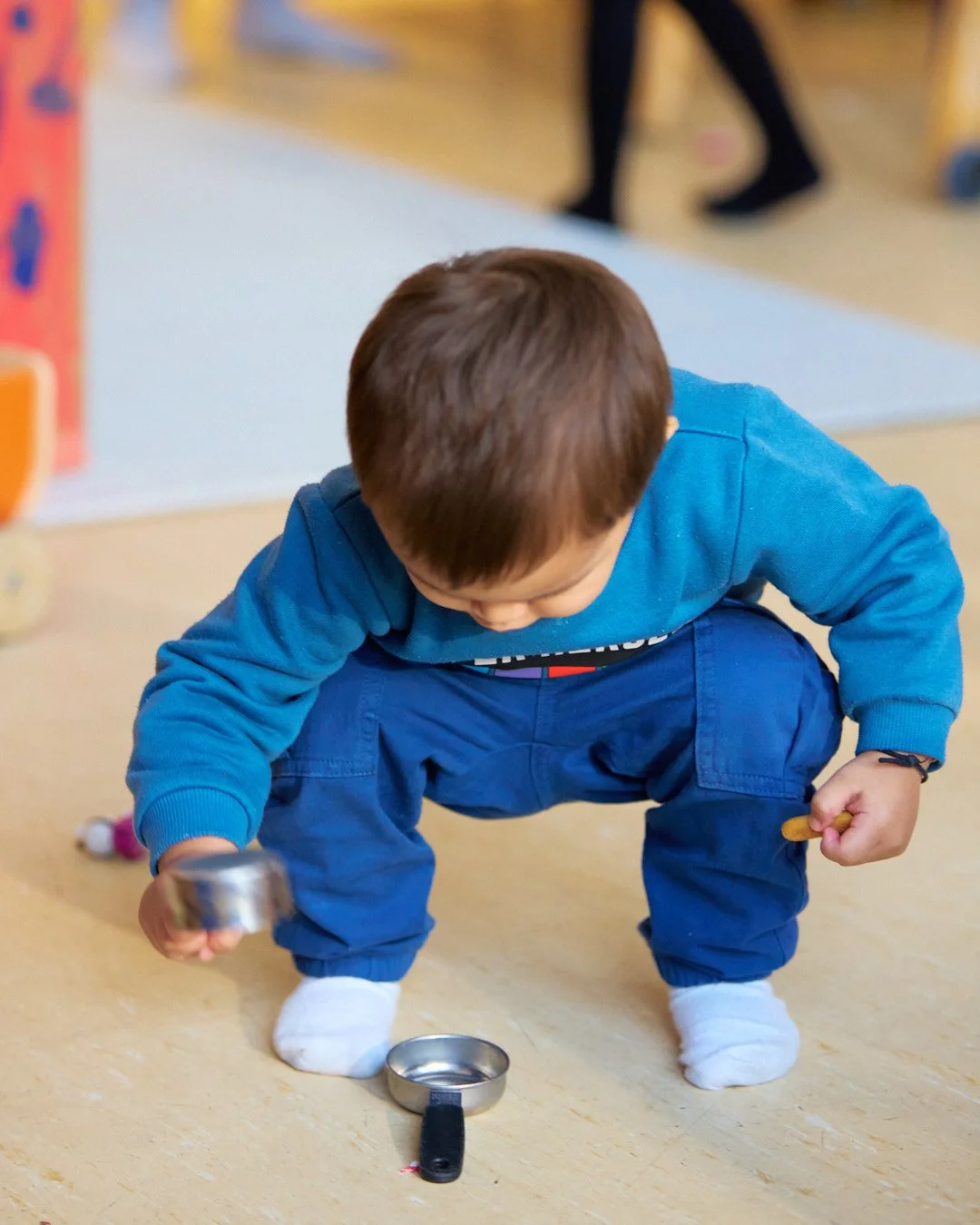 Young child crouching on the floor, playing with a small metal pan and a spoon, in an indoor setting.