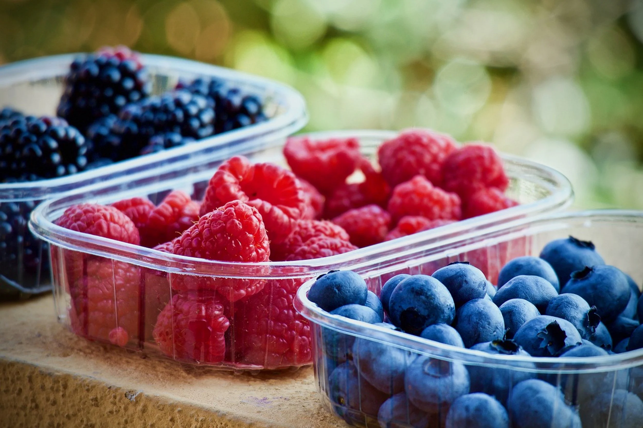 Containers of fresh blackberries, raspberries, and blueberries on a wooden surface outdoors.