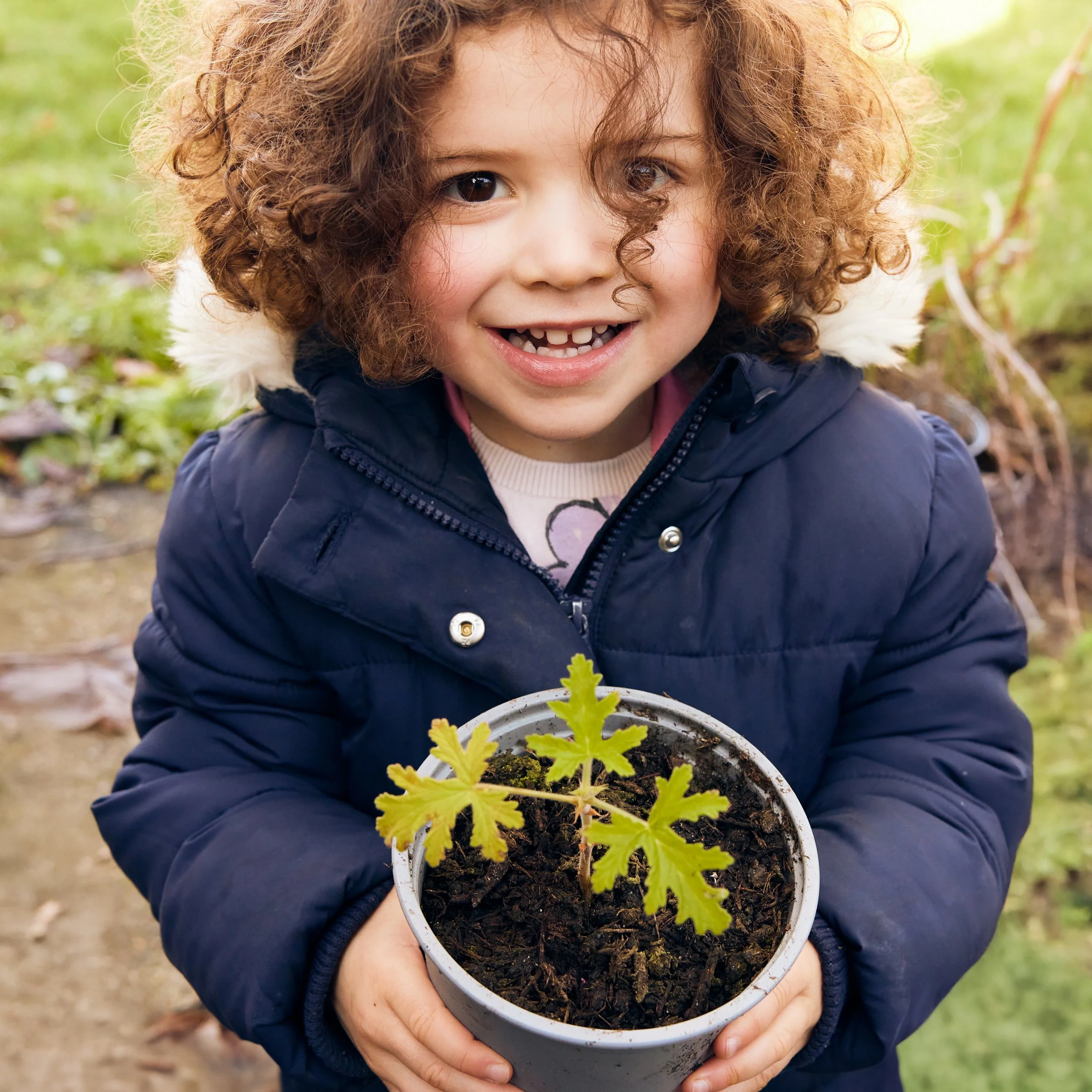 A young girl with curly hair smiling and holding a potted plant outdoors.