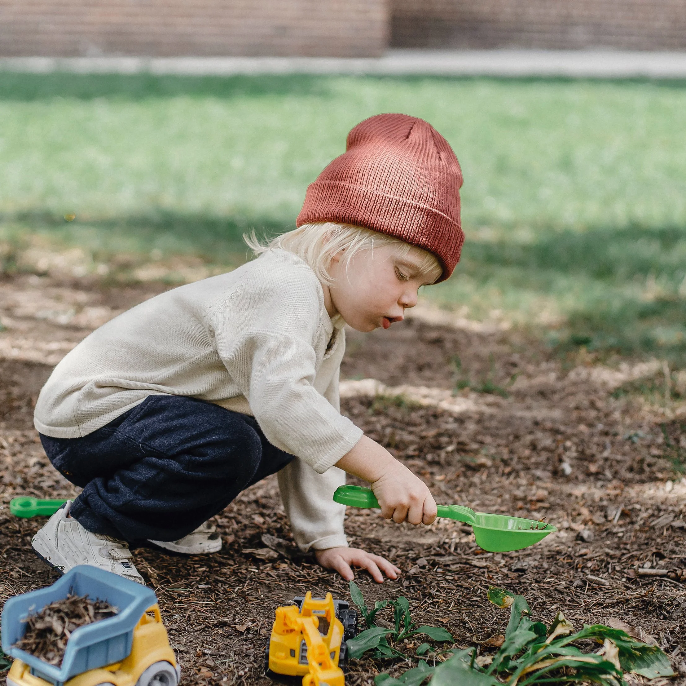 A young child with blonde hair, wearing a red beanie, a cream-colored sweater, and dark pants, squatting outdoors and using a green toy shovel to dig in the dirt surrounded by toy trucks and green leaves.