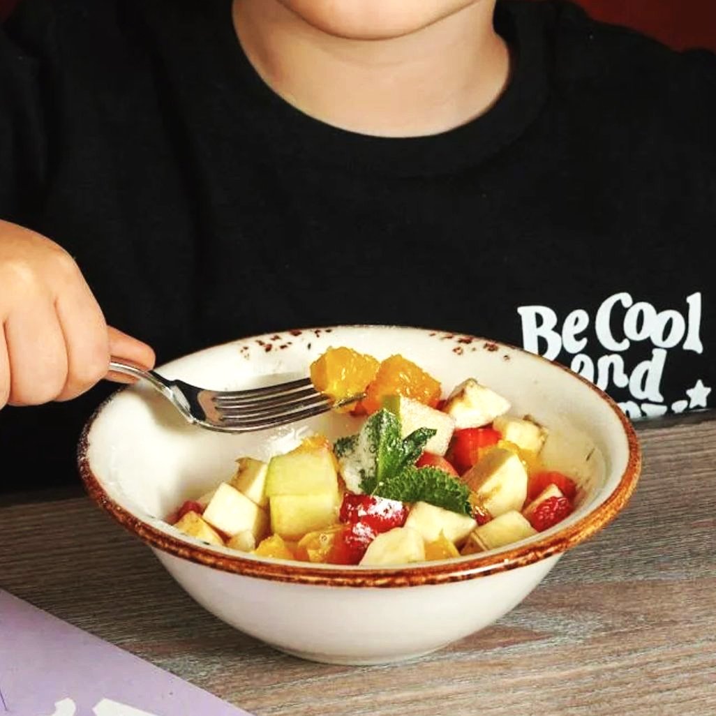 A person holding a silver fork over a bowl of fruit salad with cubes of melon, strawberries, and orange slices, garnished with mint leaves.