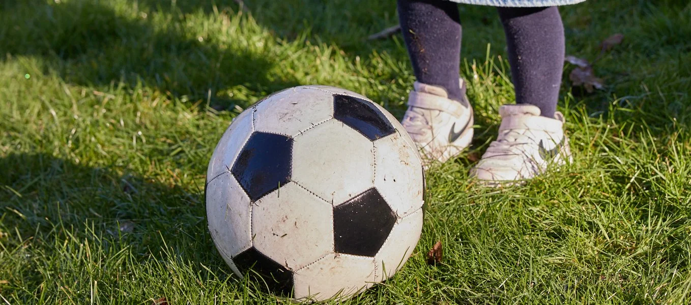 Child in white sneakers and black leggings standing next to a dirt-stained soccer ball on green grass.