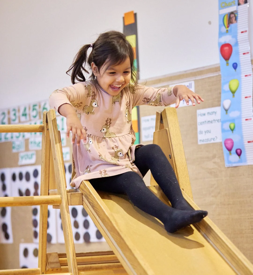 Young girl sliding down a wooden slide in a classroom, smiling with her mouth open and wearing a tan dress with reindeer print and black tights.