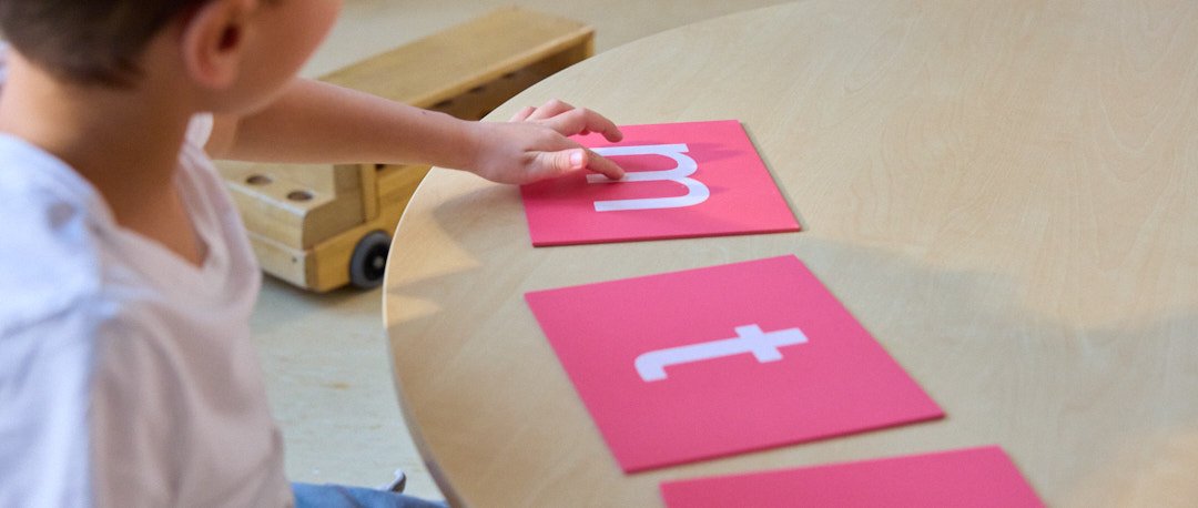 Child placing letter cards on a table, with cats of letters 't' and 'm' visible.