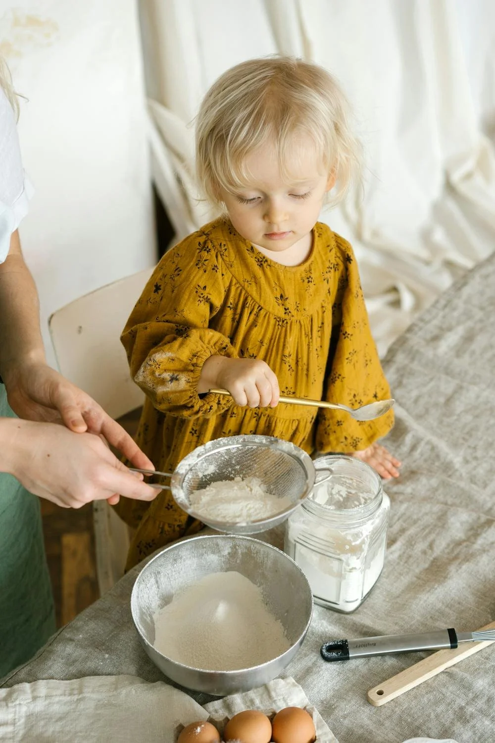 A young girl in a yellow dress sifting flour in a sieve over a bowl, with eggs and baking tools on the table.