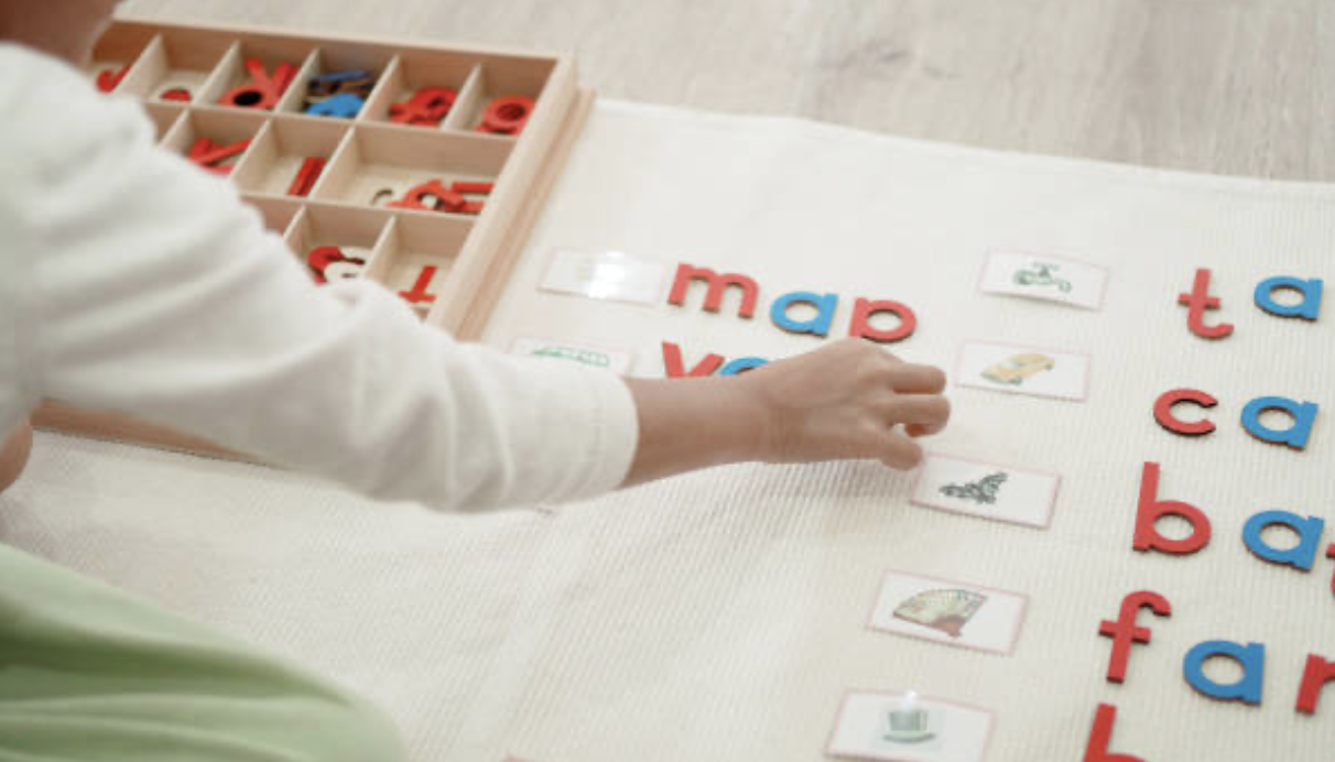 Child arranging picture cards with letters and illustrations on a table, with a box of alphabet letter tiles nearby.