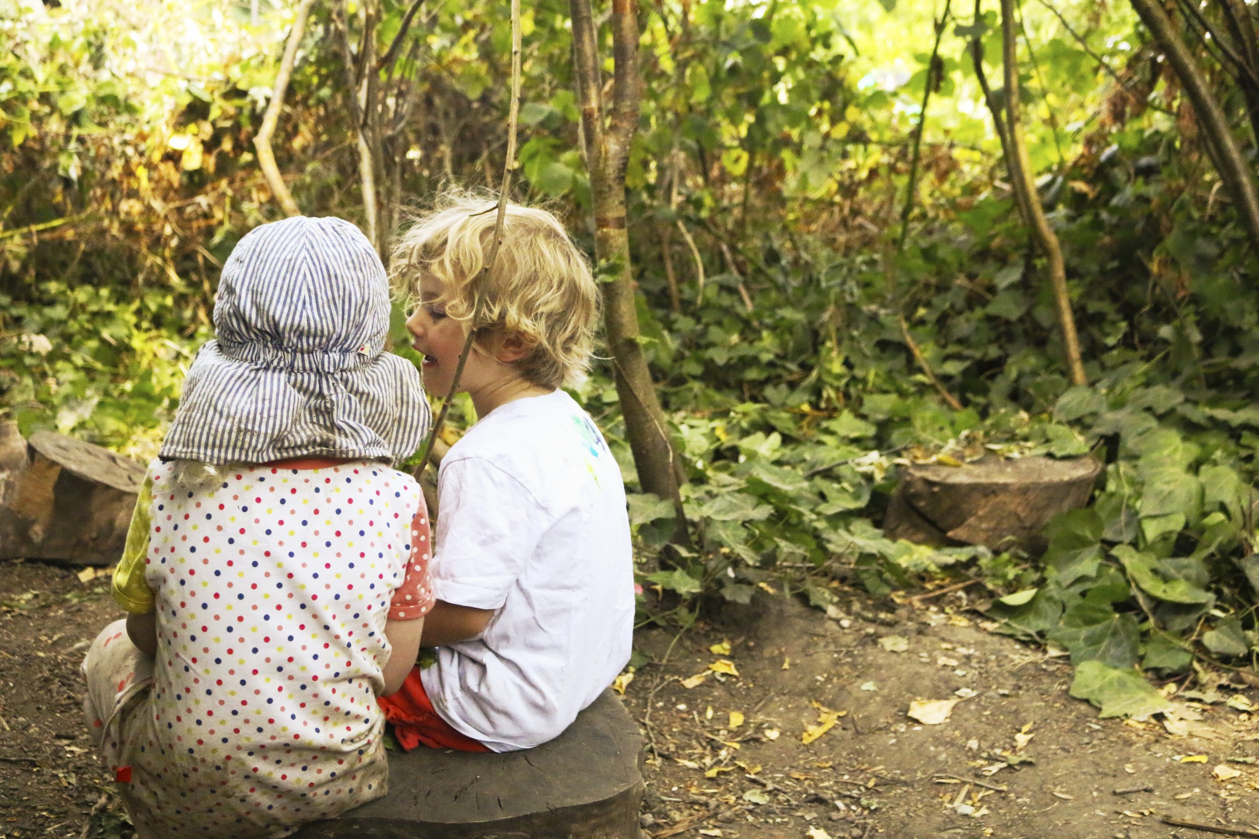 Two young children sitting on a log in a wooded area, talking and smiling at each other.