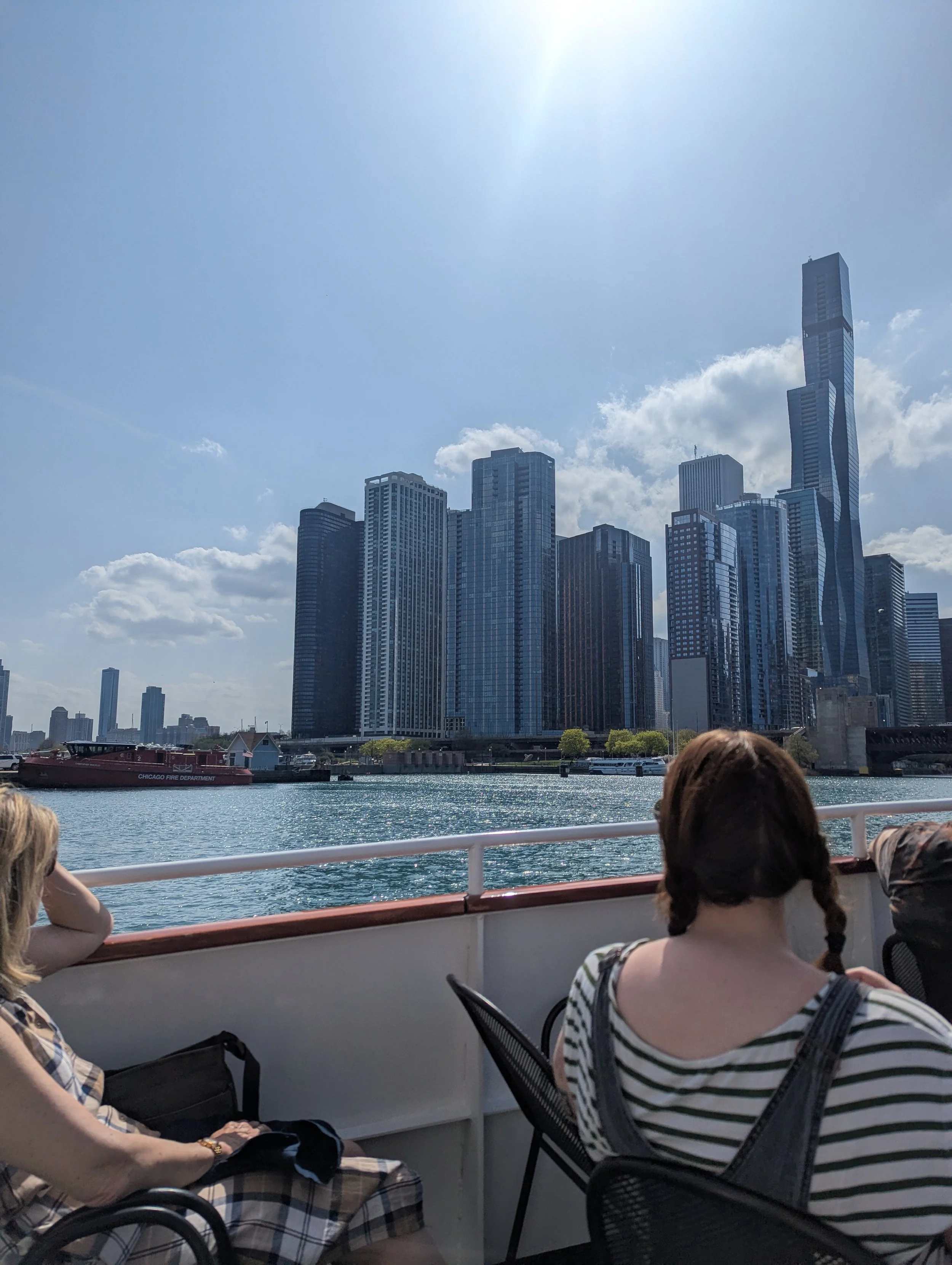 People sitting on a boat with a city skyline, including tall skyscrapers, in the background. The water is visible in the foreground, and the sky is partly cloudy with the sun shining.