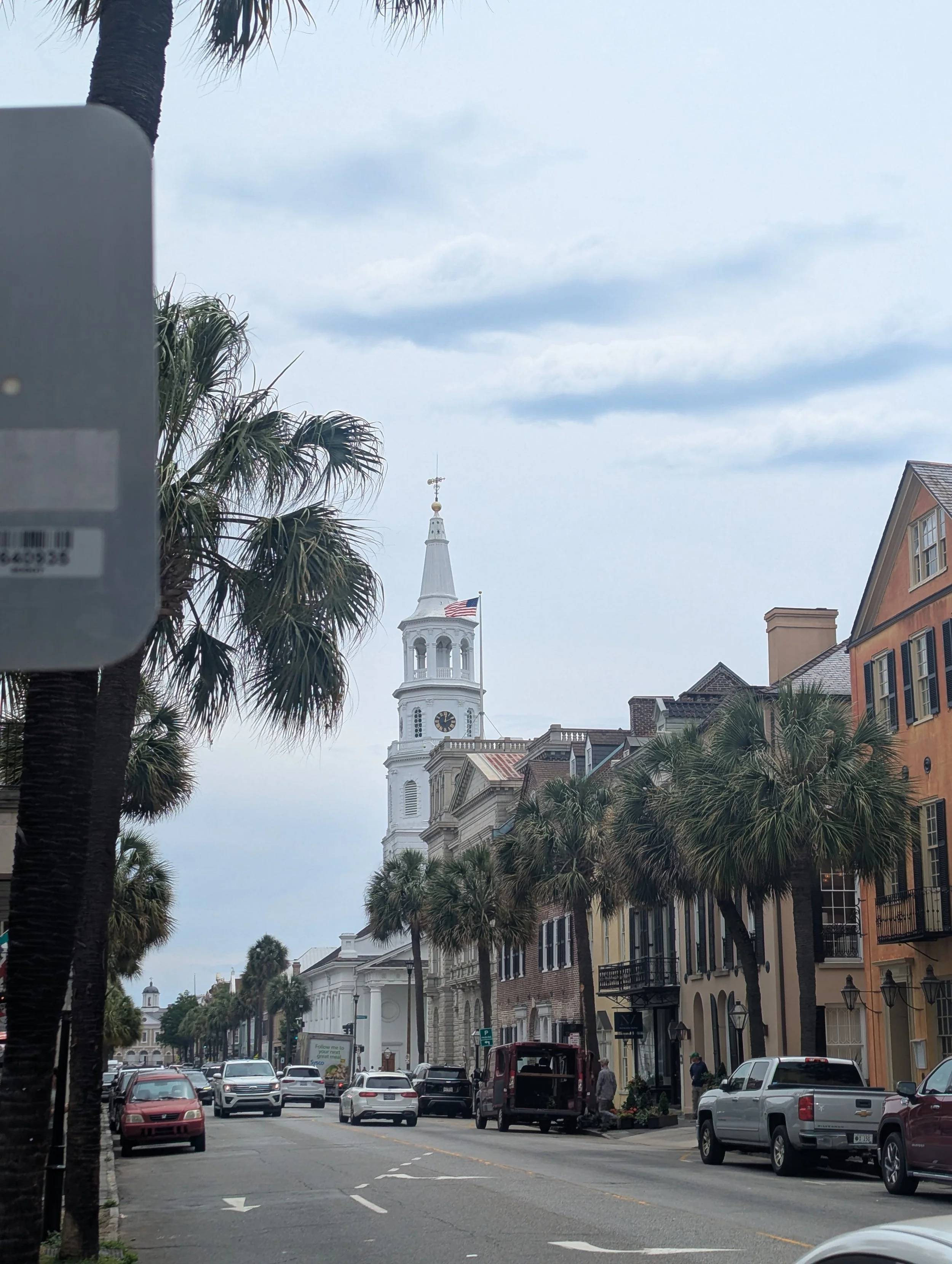 A busy city street in Charleston, SC with tall palm trees lining the sidewalk, historic buildings, and a church steeple with a clock and a flag flying on a cloudy day.