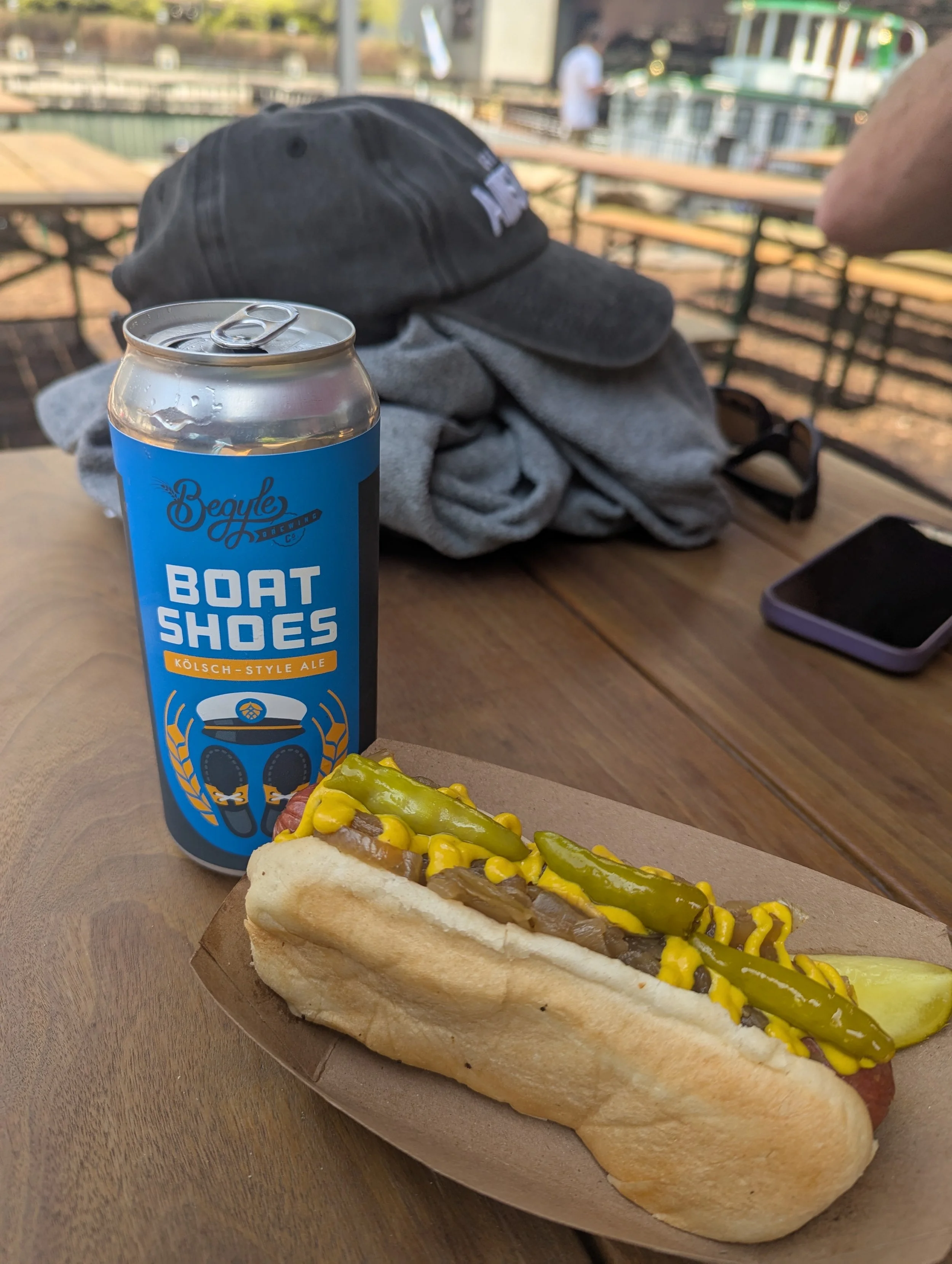 A hot dog in a bun topped with mustard, relish, and onions on a wooden tray. Behind it, there is a can of Kölsch-style ale with the branding 'Boat Shoes' and a cold beverage can with a pull-tab. The background shows an outdoor setting with a wooden table, benches, a serving boat on the water, and a person standing on a boat.