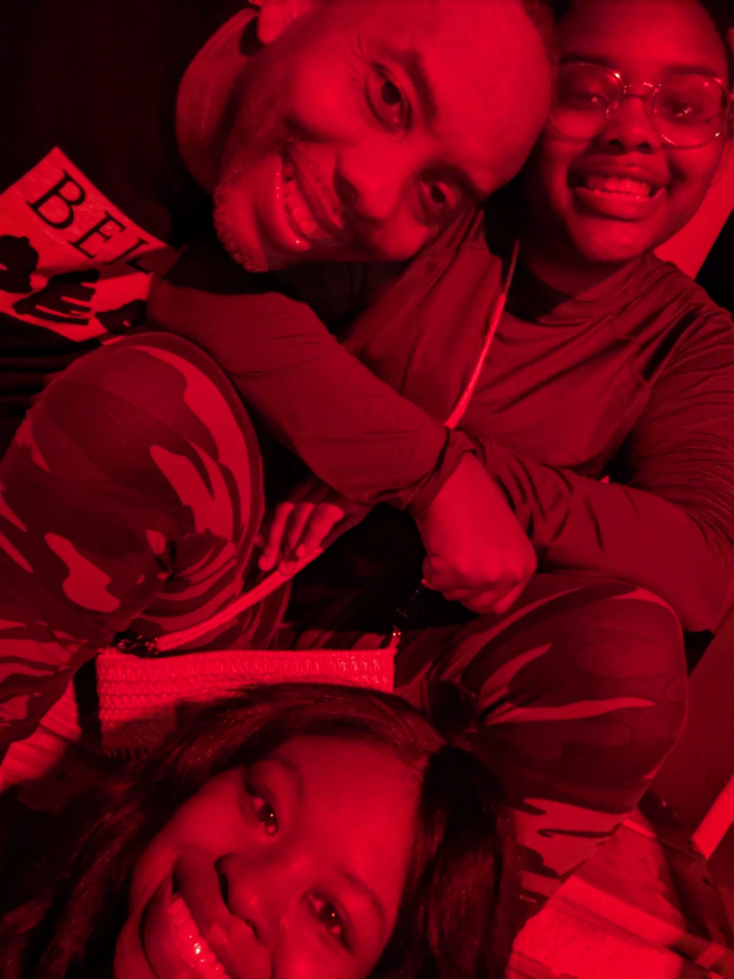 Two people smiling, one with glasses and dreadlocks, the other with makeup and a smile, in a red-lit setting.