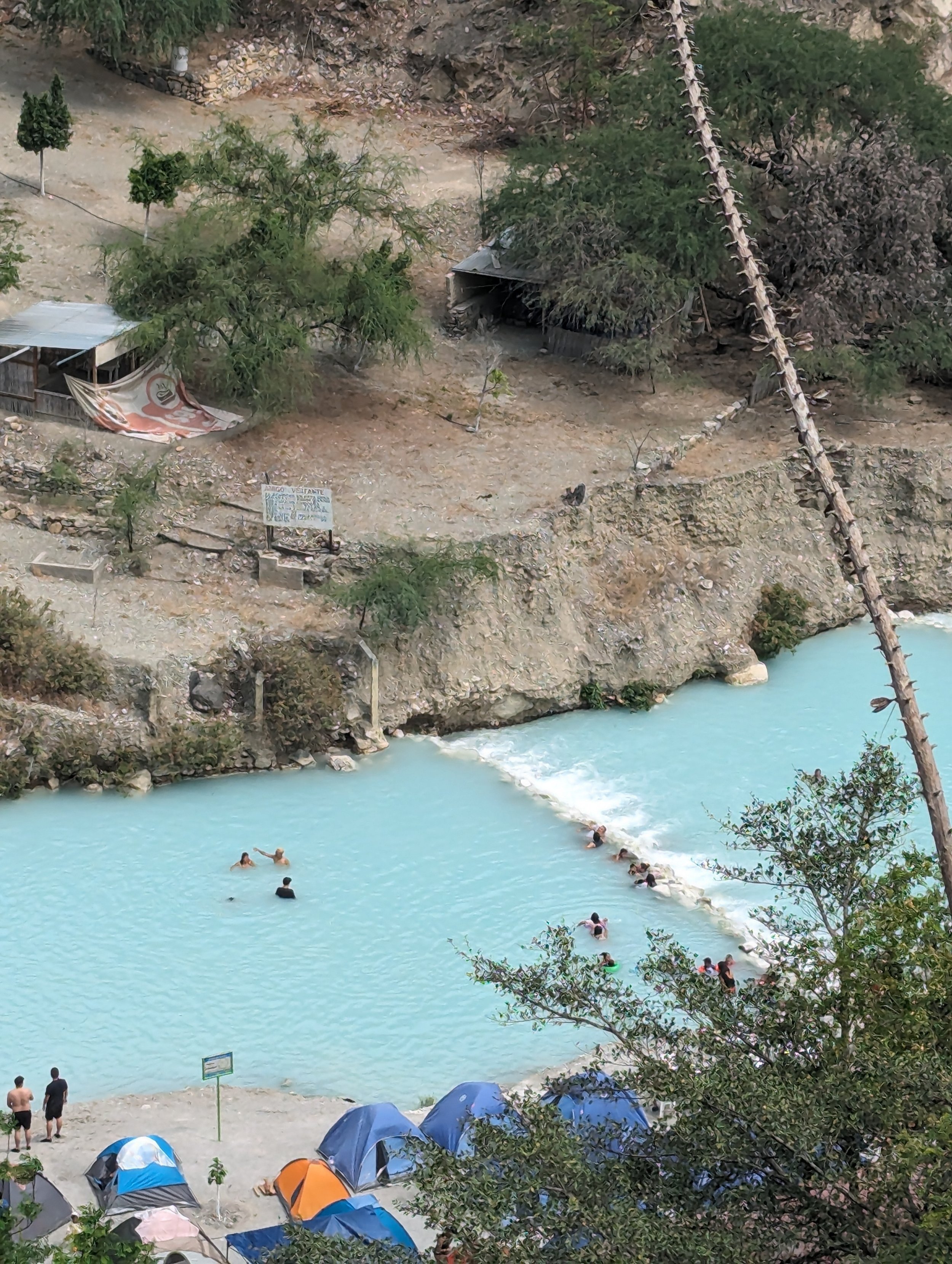 People swimming and relaxing in a turquoise river, surrounded by trees and mountains, with tents set up on the riverbank for outdoor recreation.