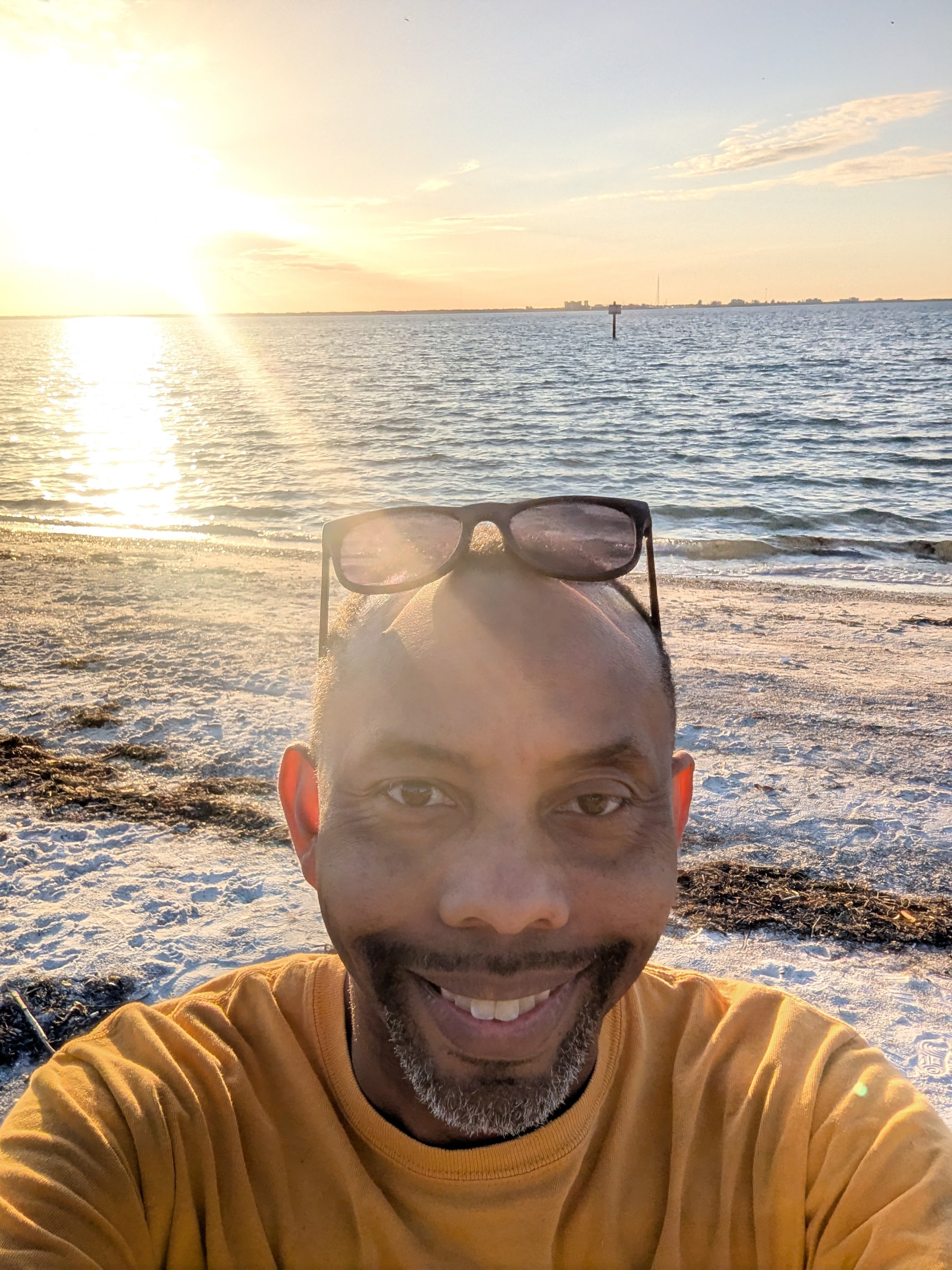 A man with a beard wearing sunglasses and a yellow shirt taking a selfie at the beach during sunset.