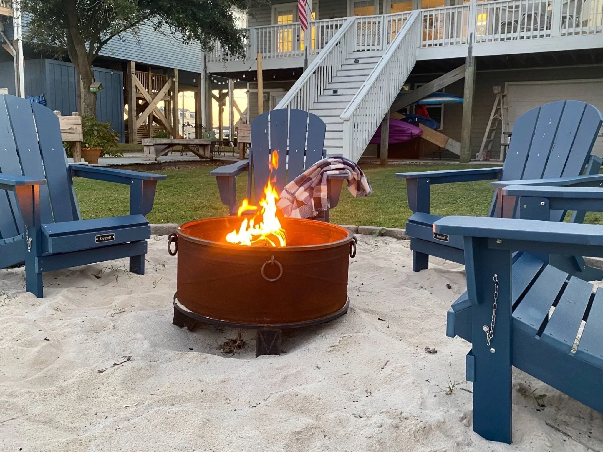 A backyard fire pit with flames surrounded by blue Adirondack chairs on sand, with a house featuring a staircase and an American flag in the background.
