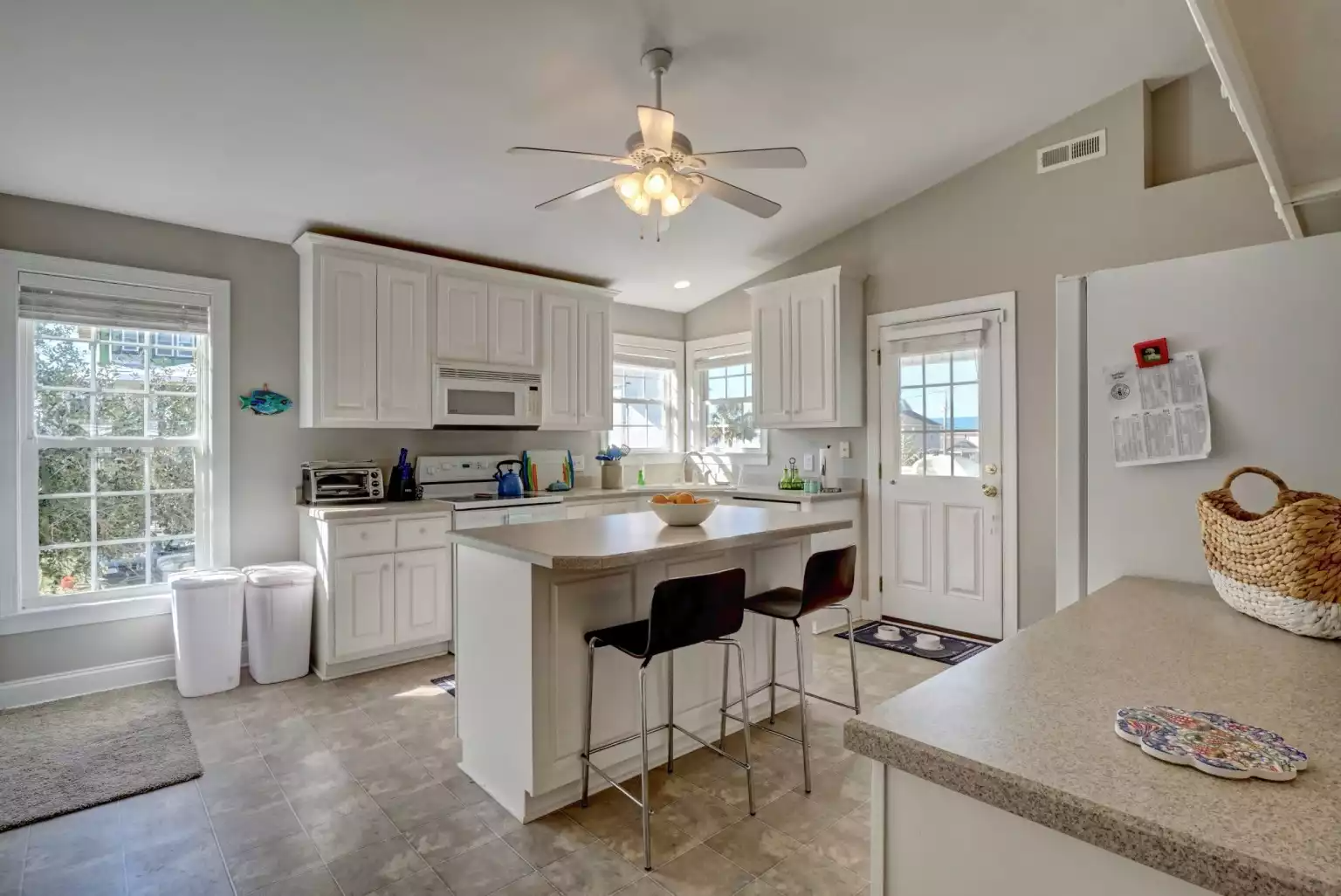 Bright kitchen with white cabinets, a central island with two black chairs, a ceiling fan with lights, large windows, a door, and tiled floor.