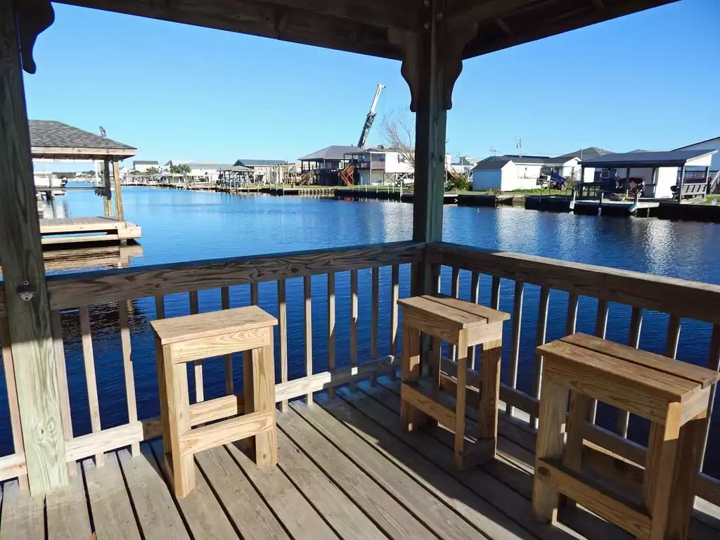View of a waterfront neighborhood seen from a wooden deck with three wooden stools, overlooking calm water and houses on the opposite side under a clear blue sky.