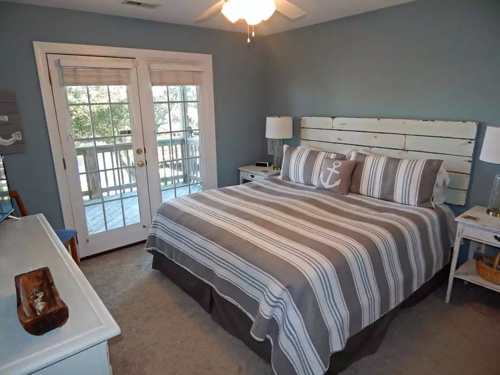 A bedroom with a large bed featuring a striped bedspread and matching pillows, a distressed white headboard, two white nightstands with lamps, a ceiling fan, and a sliding glass door leading to a balcony with trees outside.
