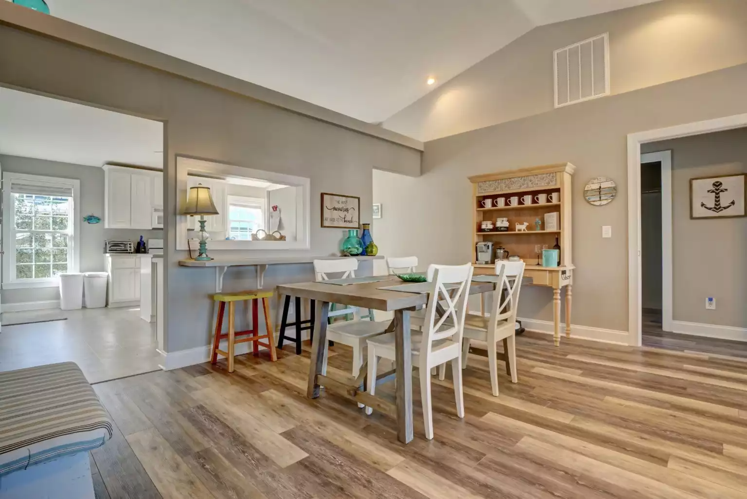 A dining room with a wooden table and six white chairs, a wooden hutch with kitchenware, and a side doorway leading to a hallway, with light-colored walls and wood-look flooring.