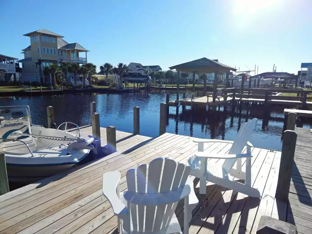 A wooden dock by a canal with two white chairs, a small boat, and houses in the background.