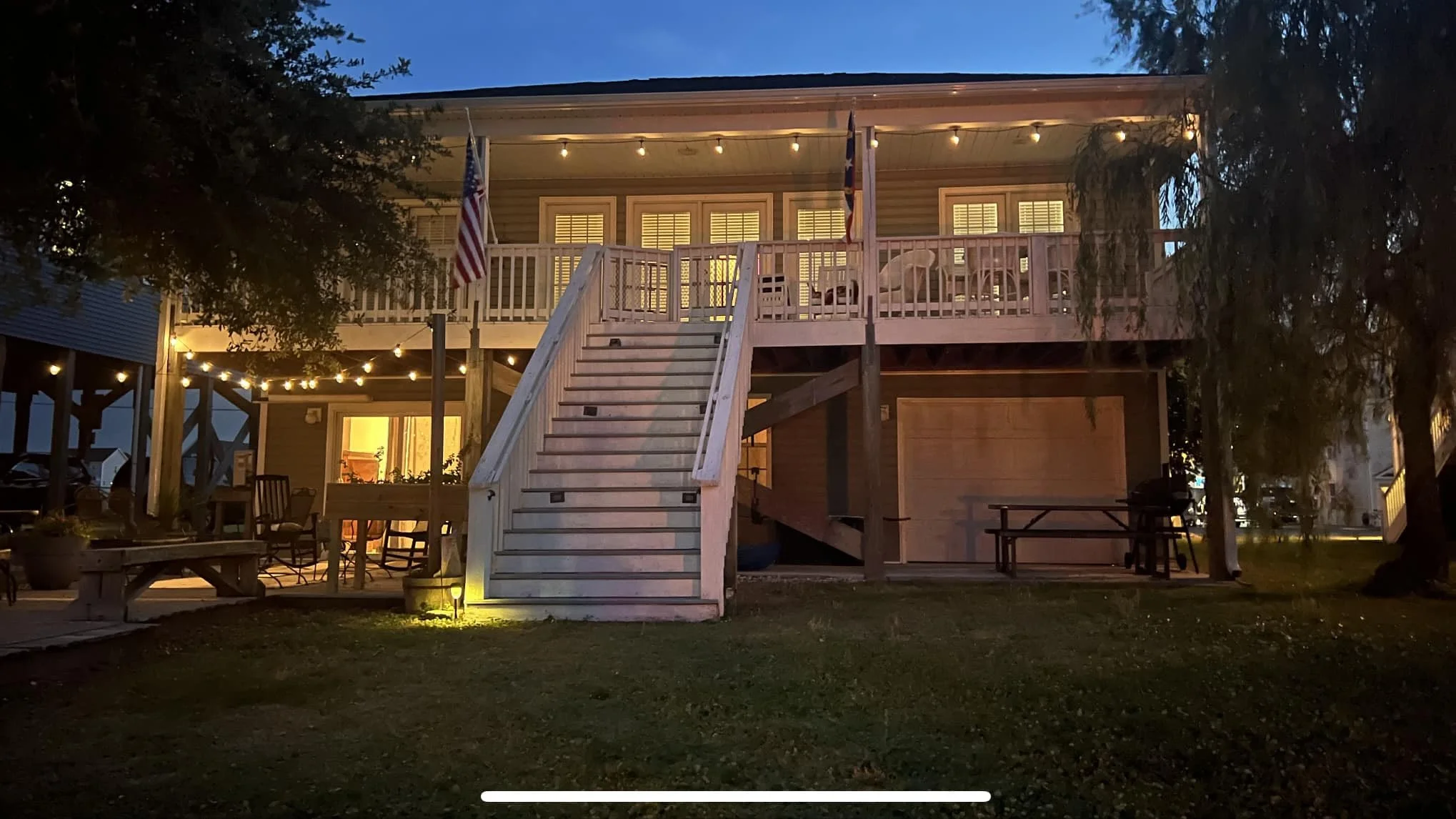 A two-story house at dusk with porch lights, American flags, and patio furniture outside. The upper deck is illuminated with string lights and has chairs and a small table.