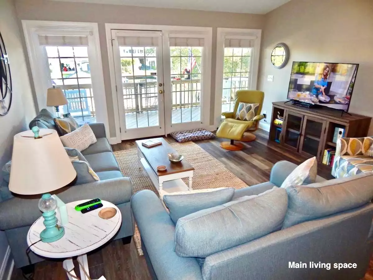 Living room with gray sofas, a yellow armchair, a TV on a wooden stand, glass doors opening to a deck, and various decorative pillows and lamps.