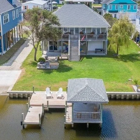 Waterfront house with a deck, stairs, and boat dock, surrounded by grass and other colorful houses.